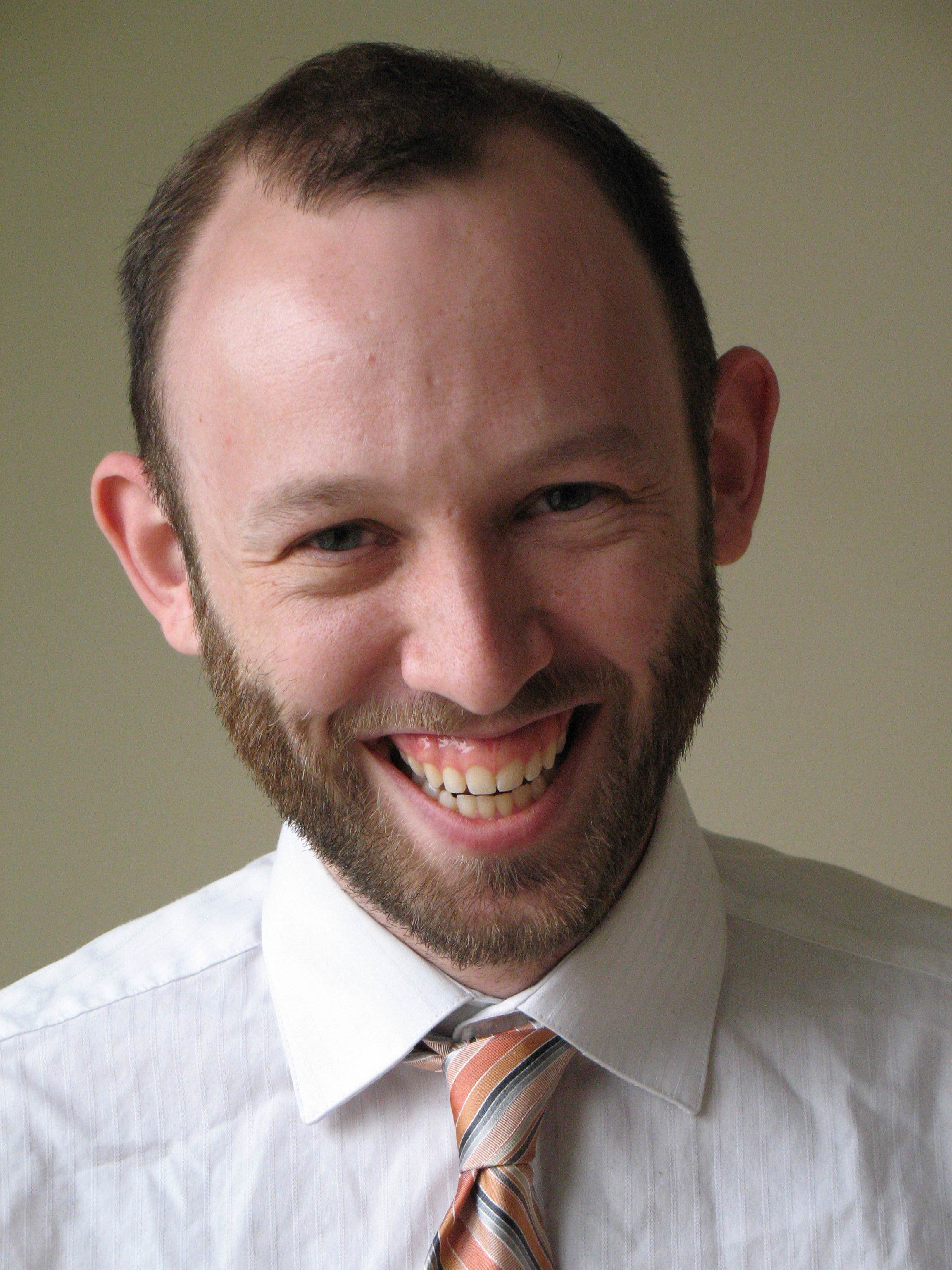 Alt text: a young white man with brown hair and a light brown clean-cut beard and mustache laughs at camera while wearing a white button down and orange and blue tie.