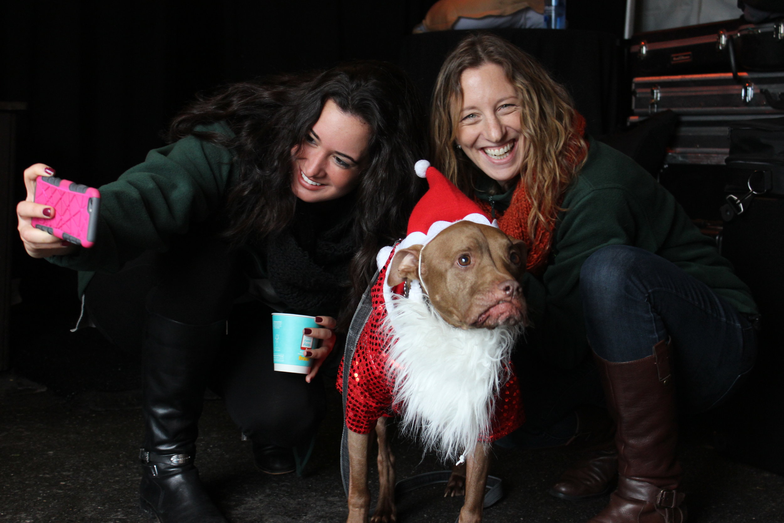 Alt text: two young white women in dark green fleece jackets smile while taking a selfie with a brown pit bull wearing a santa hat and beard costume.