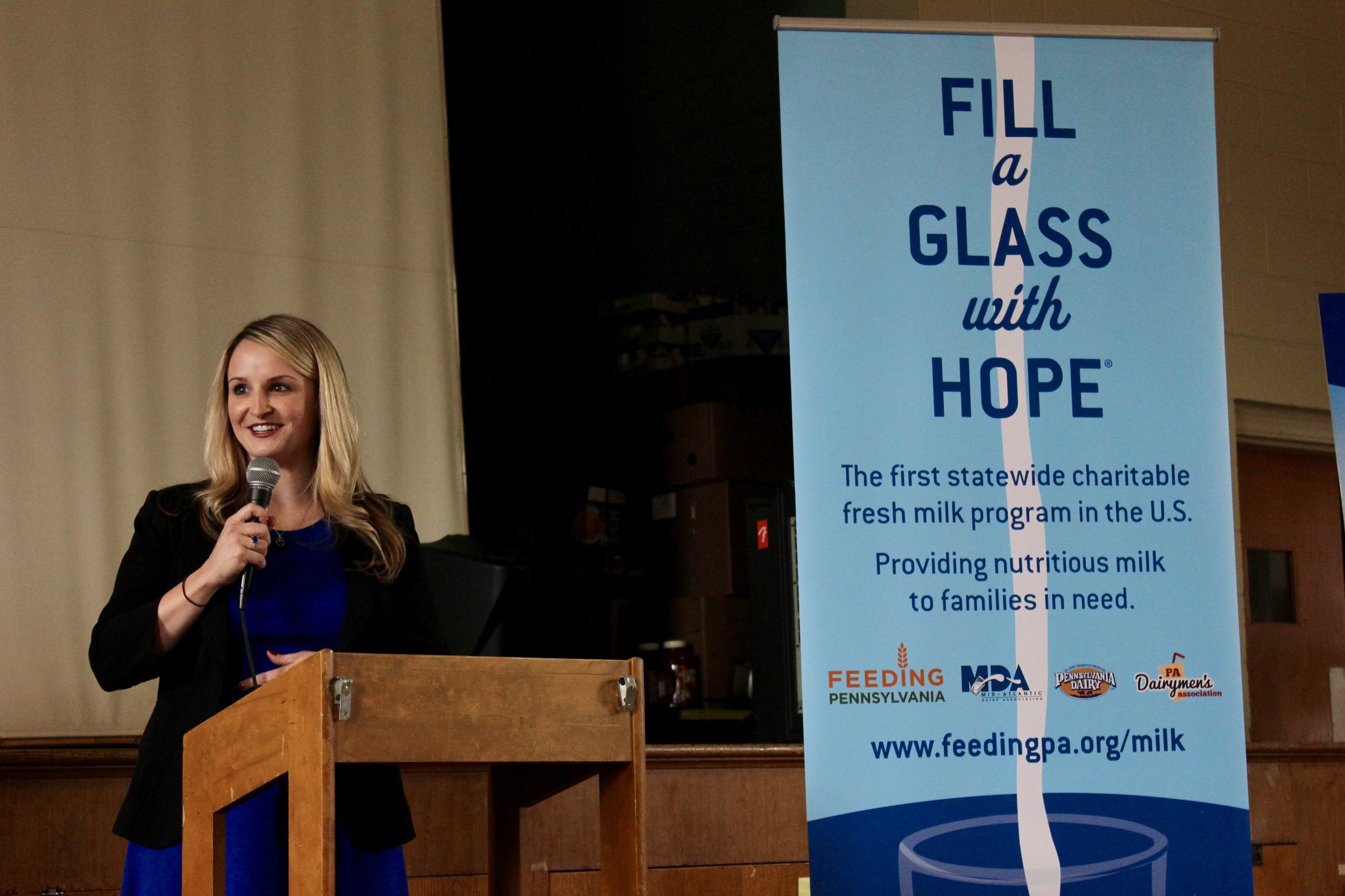 Alt text: a white woman with blonde hair holds a microphone while standing at a podium with a banner nearby that reads “fill a glass with hope”