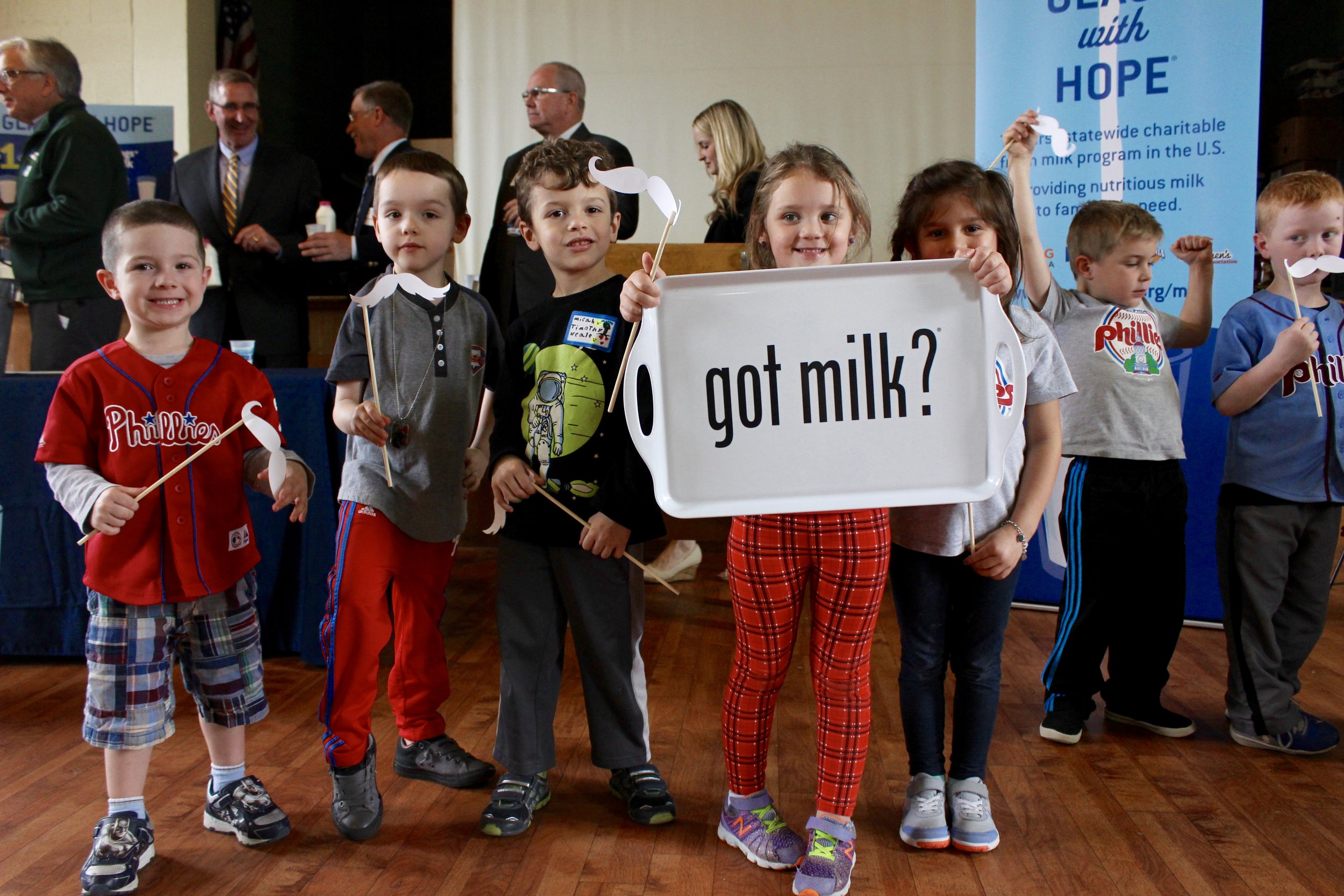 Alt text: five little kids stand together while holding photobooth props and smiling towards camera. One little girl with light brown hair and bright red leggings holds a sign that reads “got milk?”
