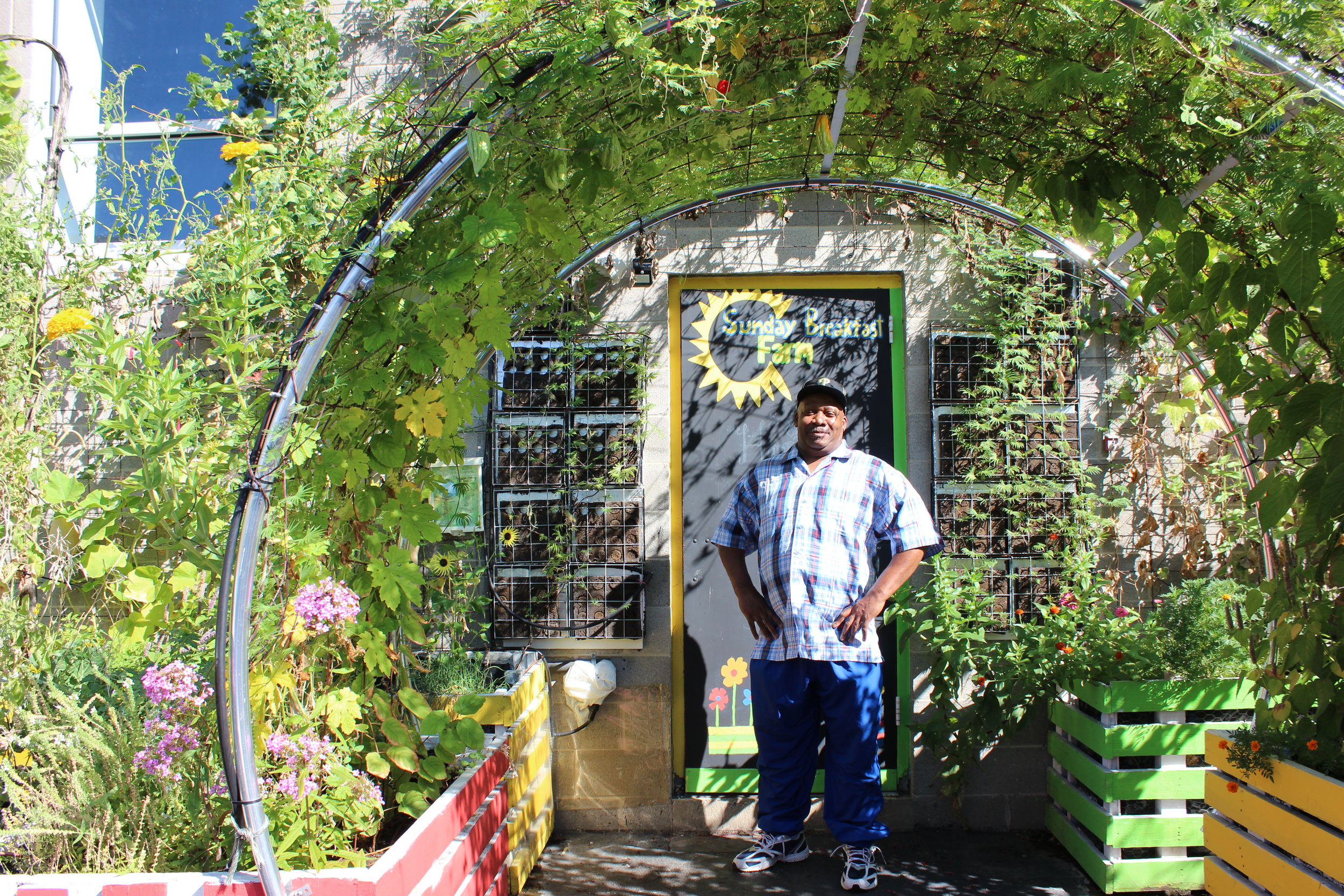 alt text: a sunny wideshot of a black man wearing a baseball hat, checkered button down, and blue jeans stands smiling proudly at the camera. An archway of lush flowers and leaves arches over him.
