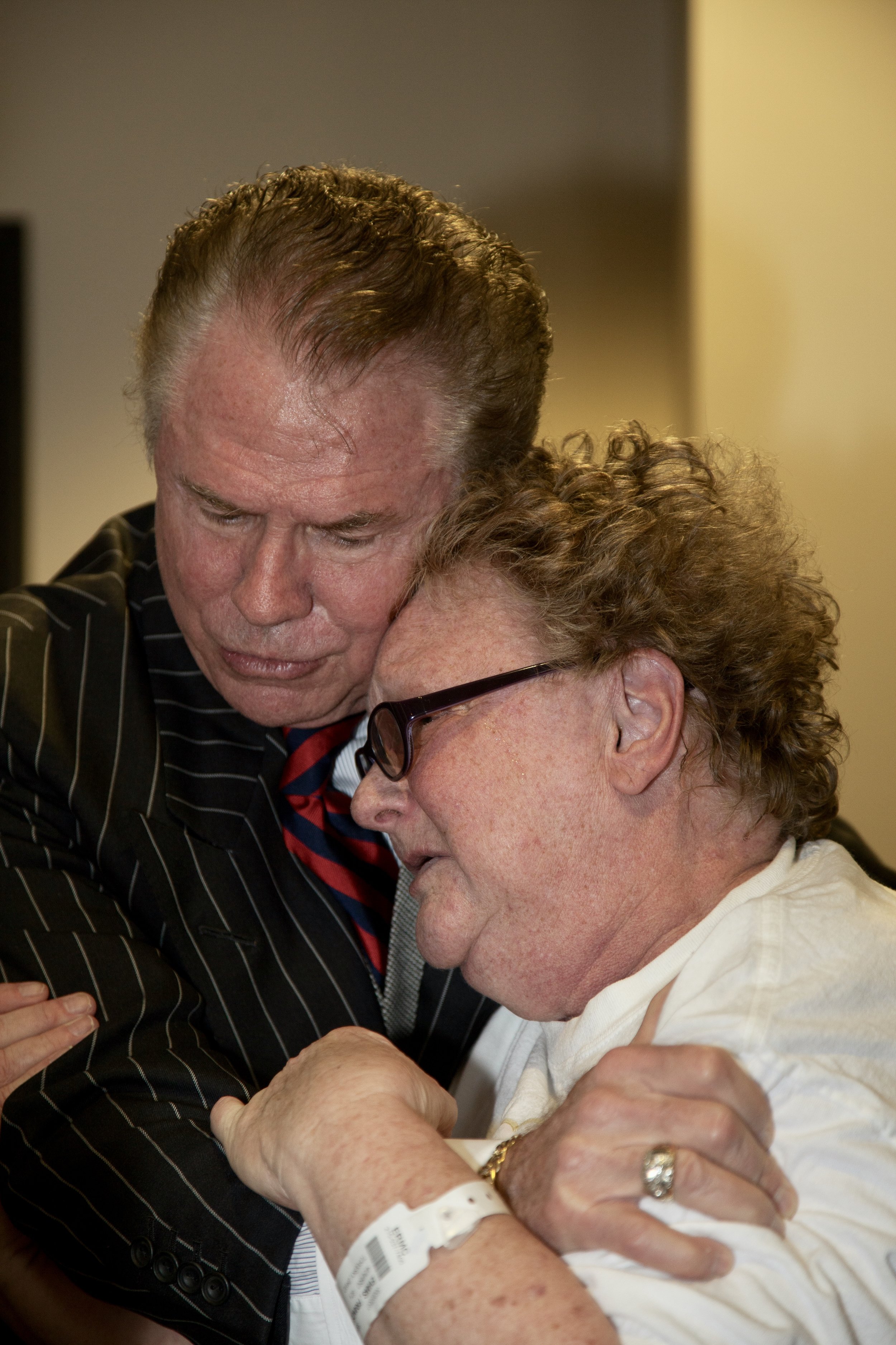 alt text: an older white man with a receding hairline and wearing a pinstripe suit consoles an older white woman wearing black thick-rimmed glasses as well as a hospital band on her wrist