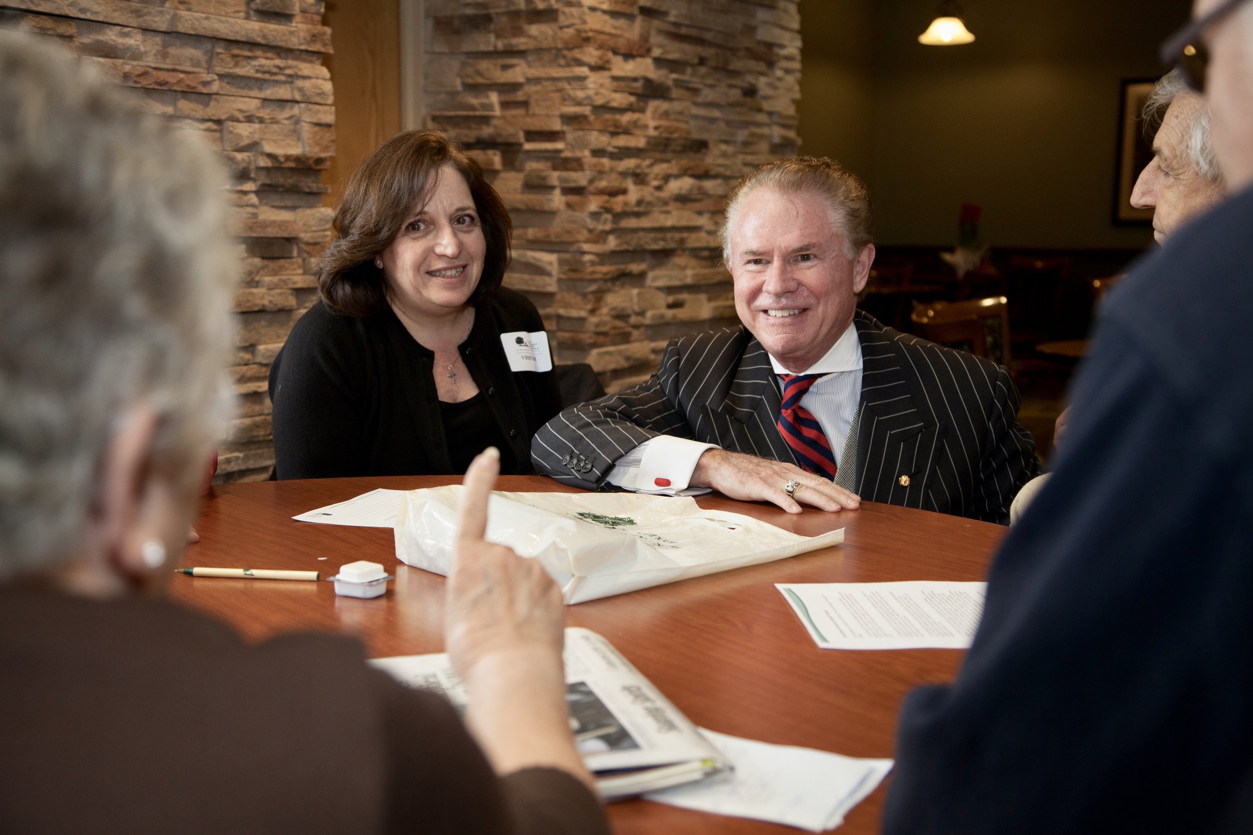 Alt text: an older white man with a receding hairline and wearing a pinstripe suit crouches down at a table and smiles at the other people sitting at the table, including a middle-aged white woman wit