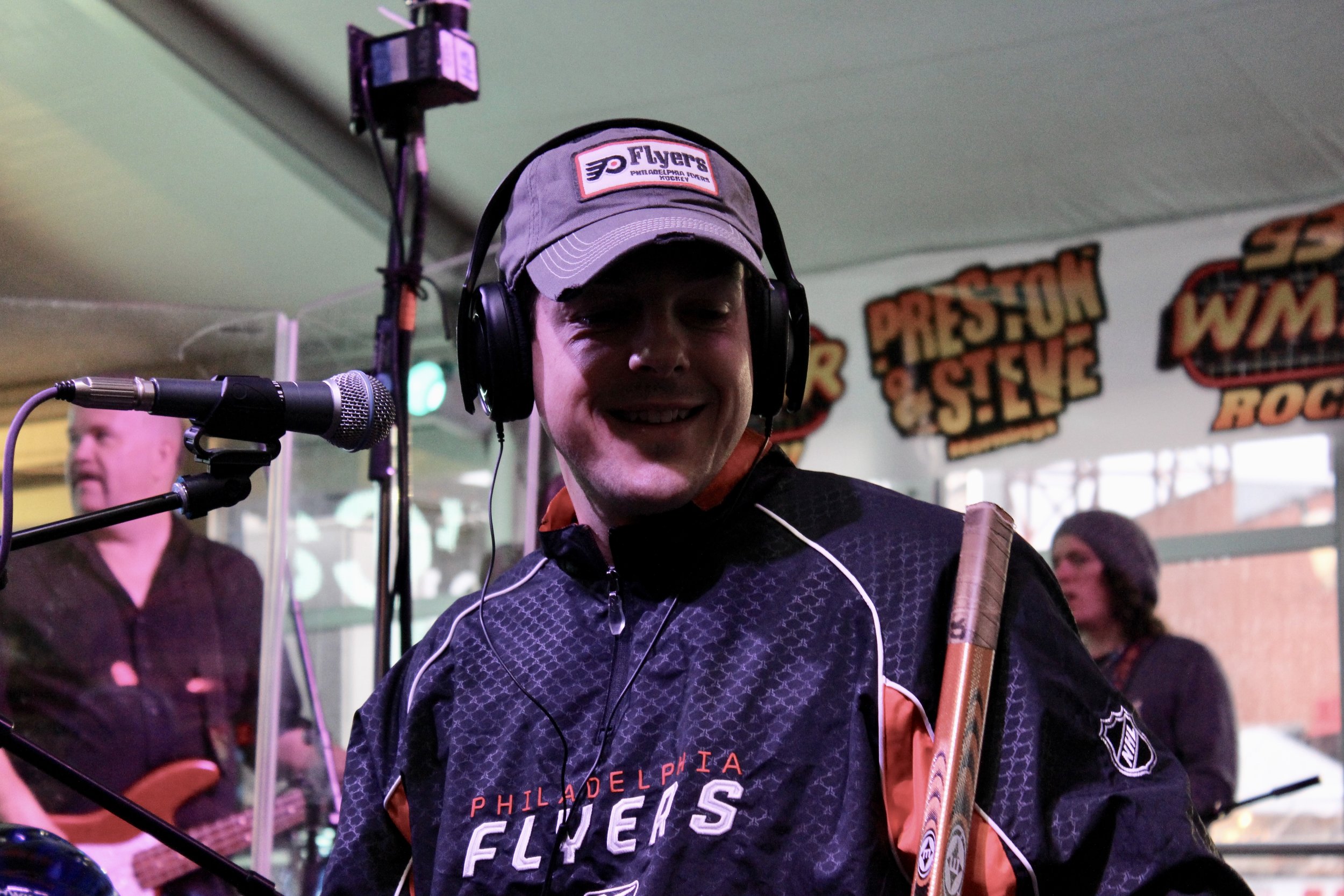 Alt text: philadelphia flyers hockey player has headphones on and sits at a microphone while holding his hockey stick to talk on a radio show.