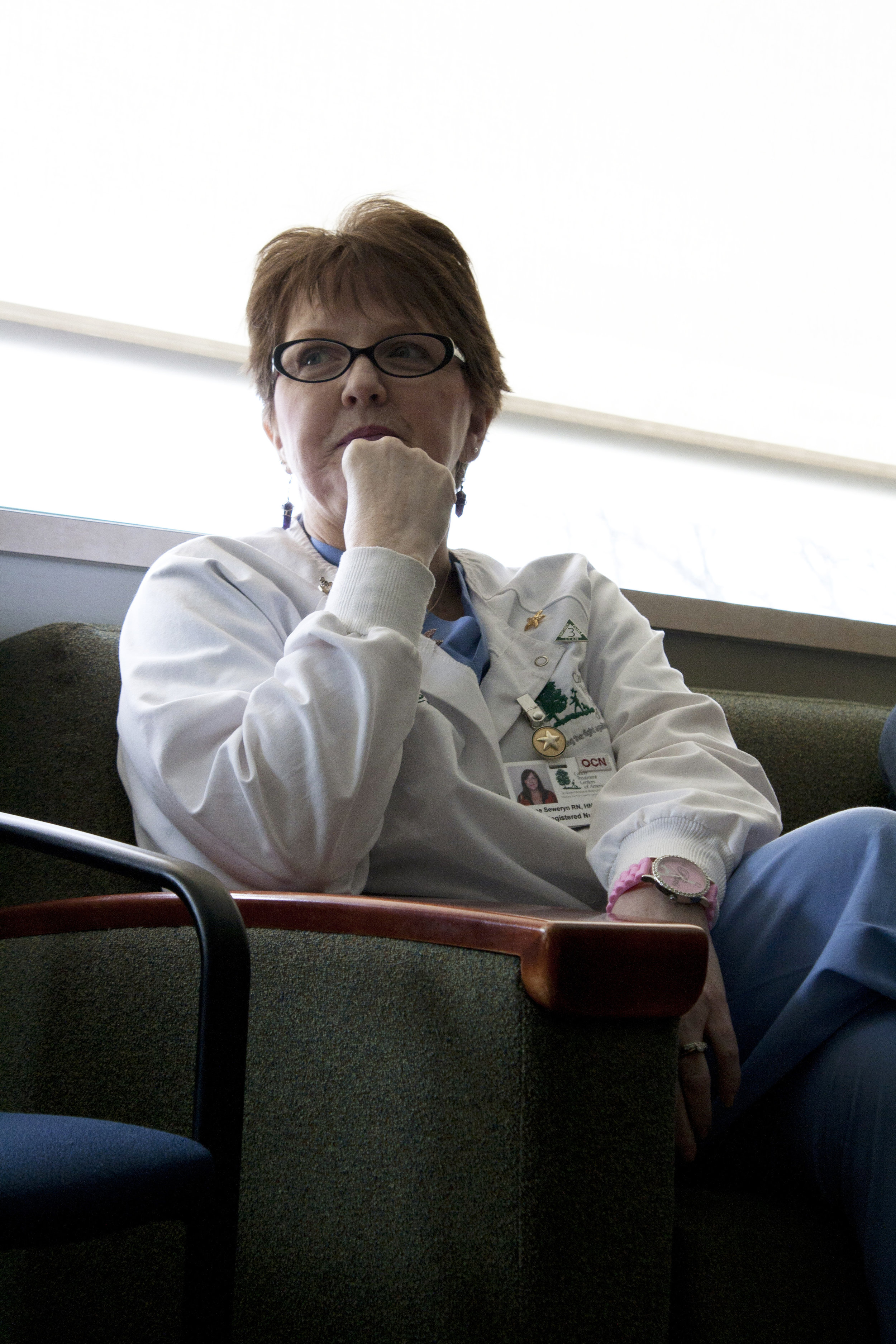 Alt text: a middle-aged white woman with short brown hair and oval, thick-rimmed glasses looks pensively off camera while sitting in a green chair. She’s wearing blue scrubs and a nurse’s lab coat wit