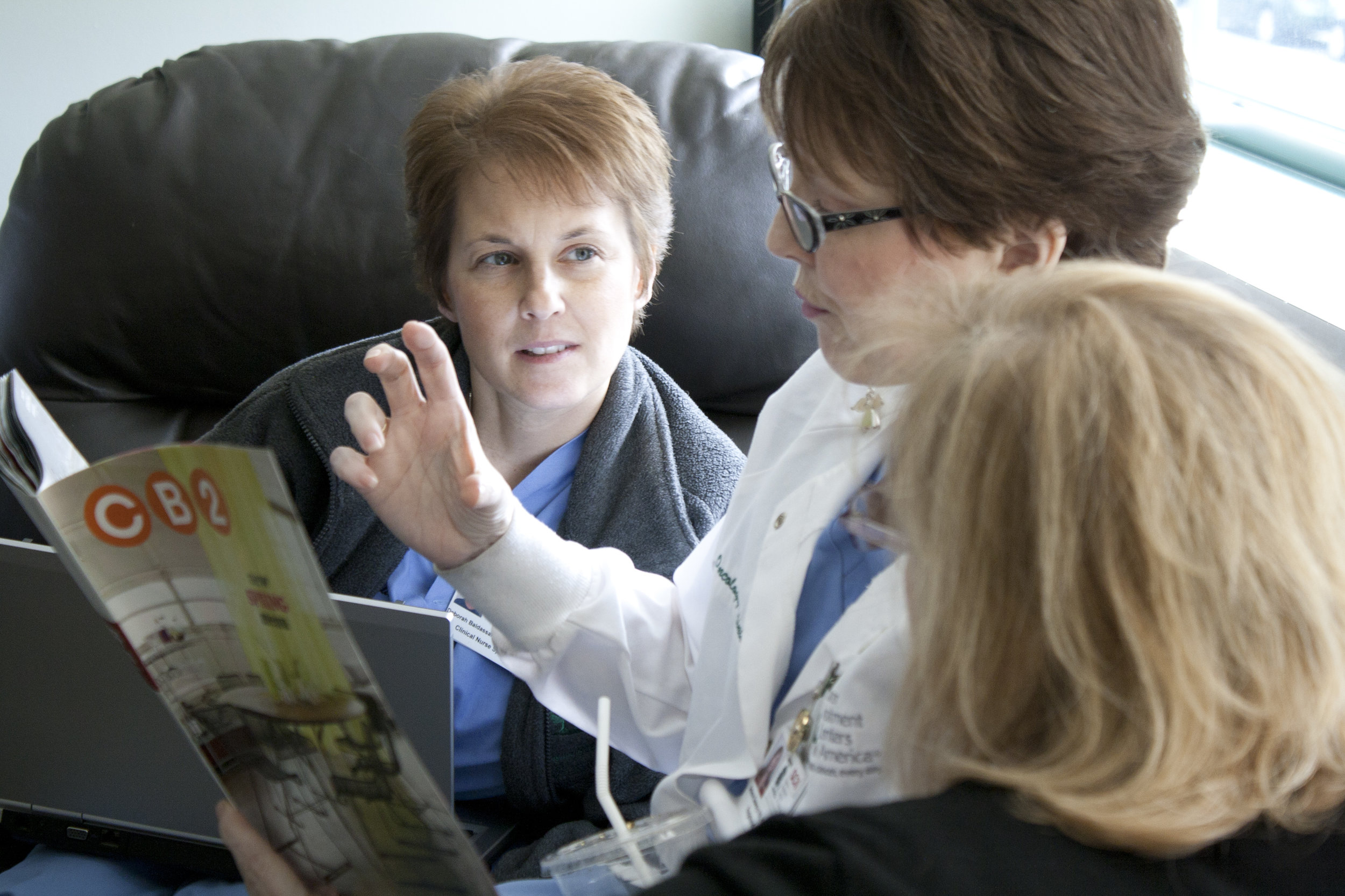 Alt text: a middle-aged white woman with short brown hair and oval, thick-rimmed glasses sits between two other young women who are looking at her as she reads from a magazine in her hands.