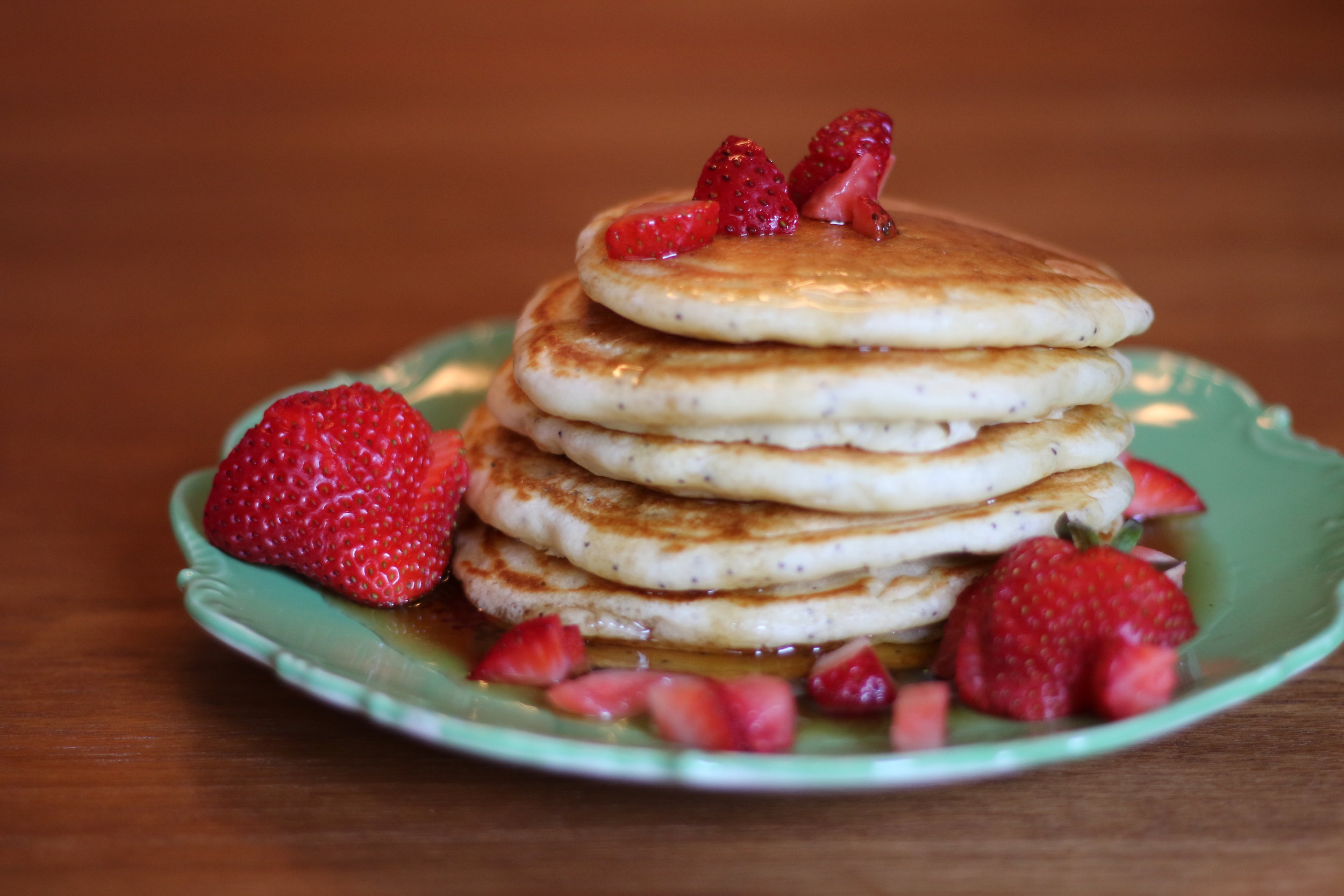 Alt text: a stack of 5 pancakes sit on a pretty green plate, covered in syrup and cut strawberries