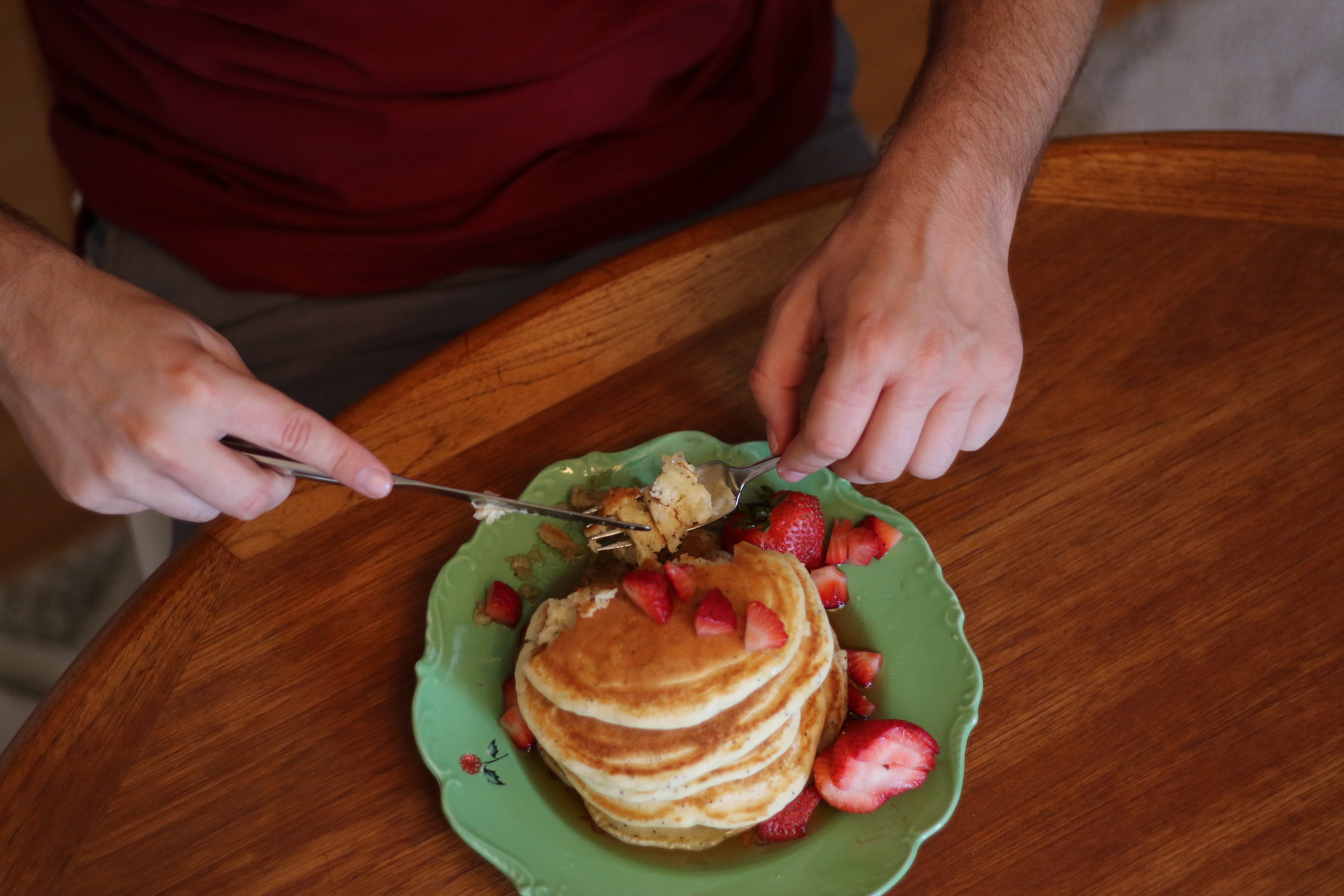 Alt text: a bird’s eye view of white hands holding silverware and cutting into a stack of 5 pancakes sit on a pretty green plate, covered in syrup and cut strawberries