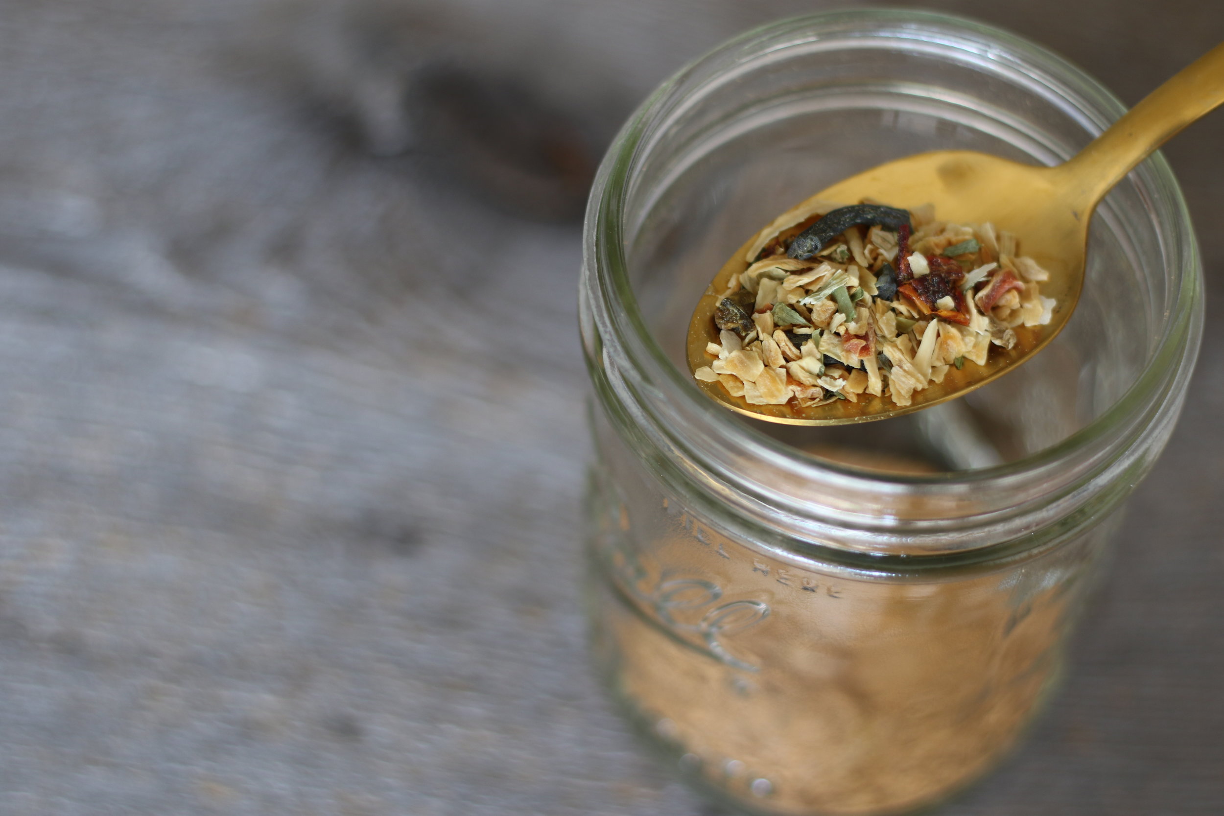 Alt text: a small mason jar of spices sites on a wooden tabletop. At the top of the image, you can see a gold spoon pulling out of the jar with colorful spices in it.