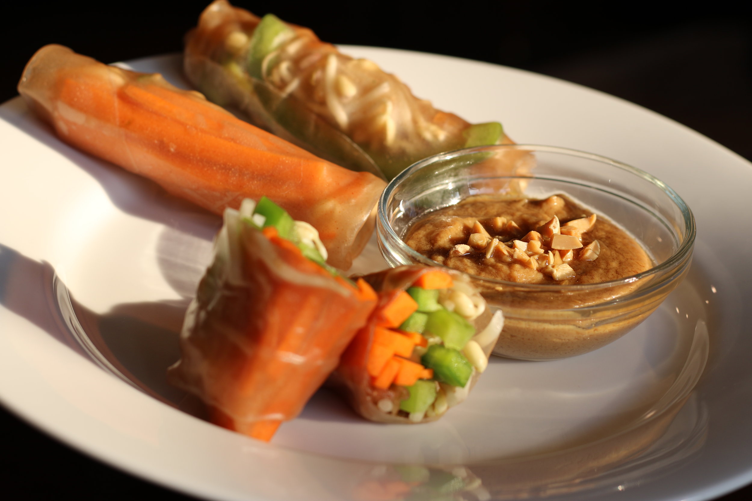 Alt text: a white plate holds vegetable spring rolls in rice wraps along with a small dish of peanut butter sauce. The light is very moody with sharp shadows.