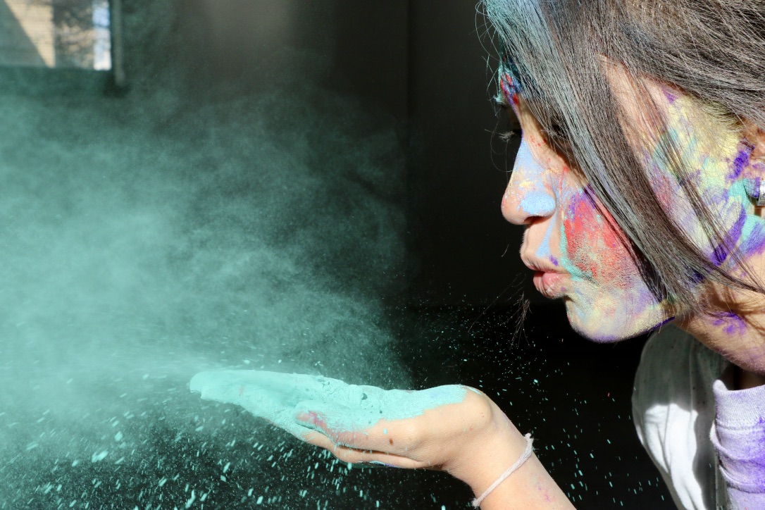 alt text: a closeup of an indian american teenage girl with brown bangs covering her eyes and colored powders swirling across her face as she blows into her hand to scatter vibrant blue powder during 