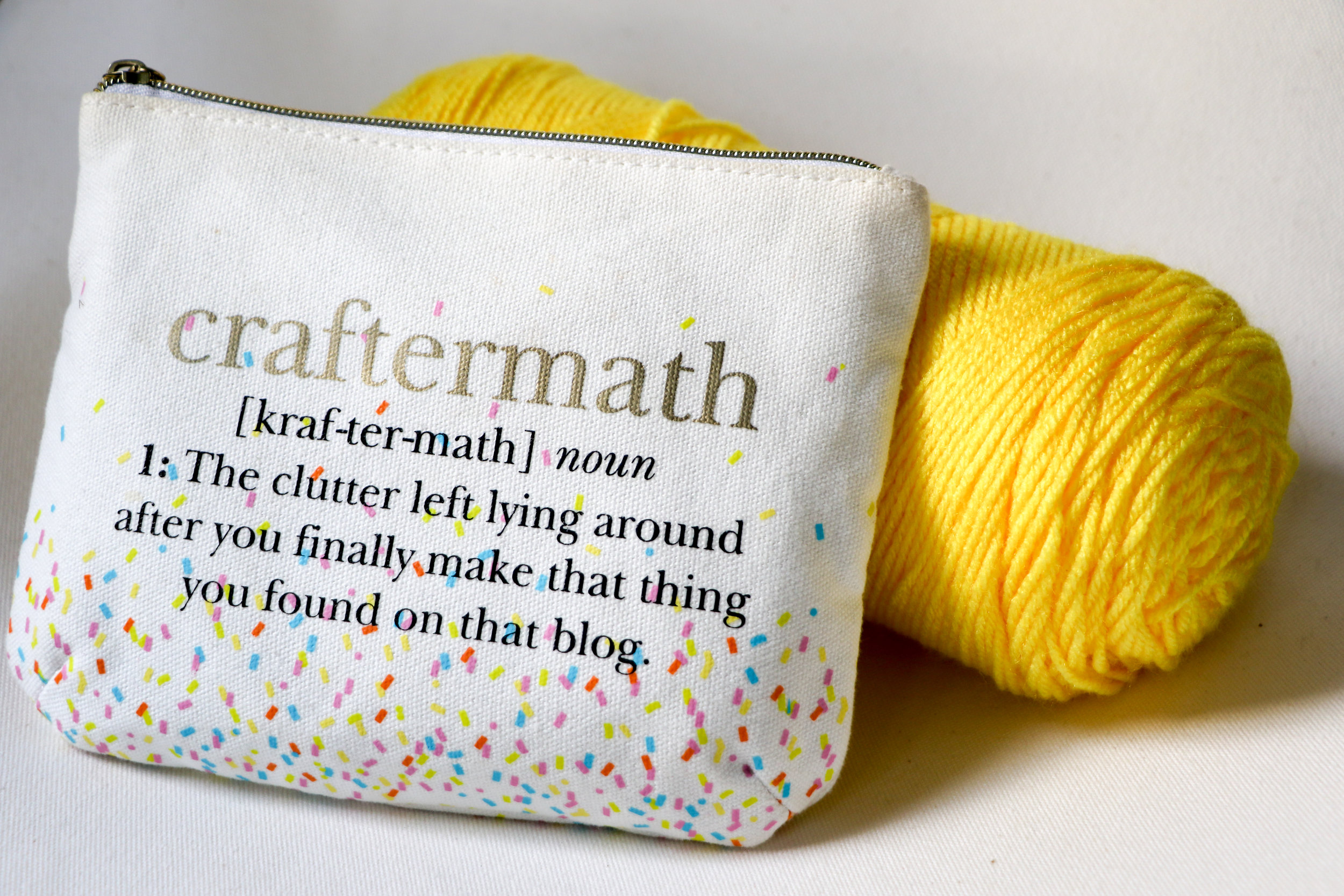 Alt text: a canvas zipper bag with confetti decals and text that reads “craftermath” and a definition for the word leans against a bright yellow ball of yarn.