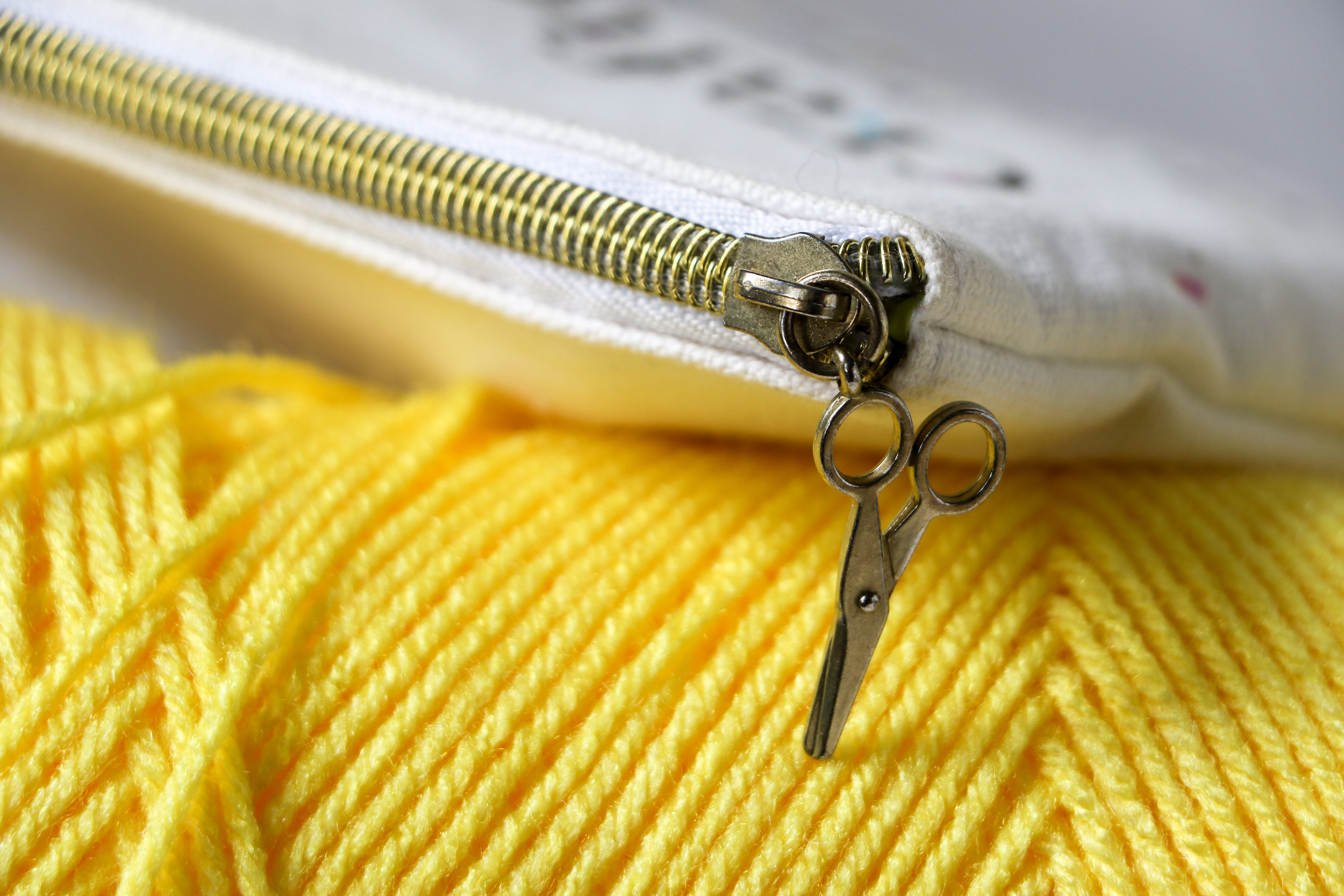 Alt text: a closeup of a brass zipper with a charm that looks like a pair of scissors hands off a canvas bag and leans against a bright yellow ball of yarn.