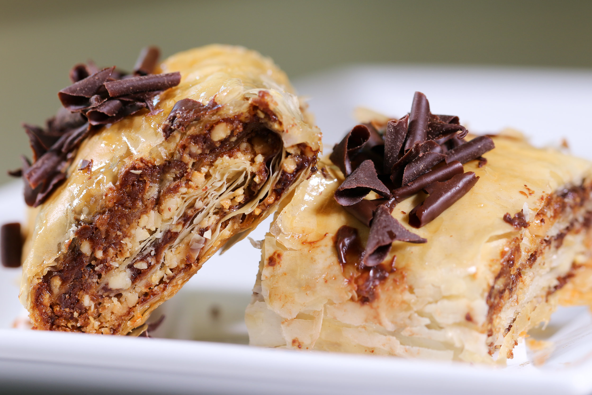 Alt text: a close up of two pieces of baklava that rest against one another and include layers of flaky pastry, nuts, brown sugar, and flakes of chocolate