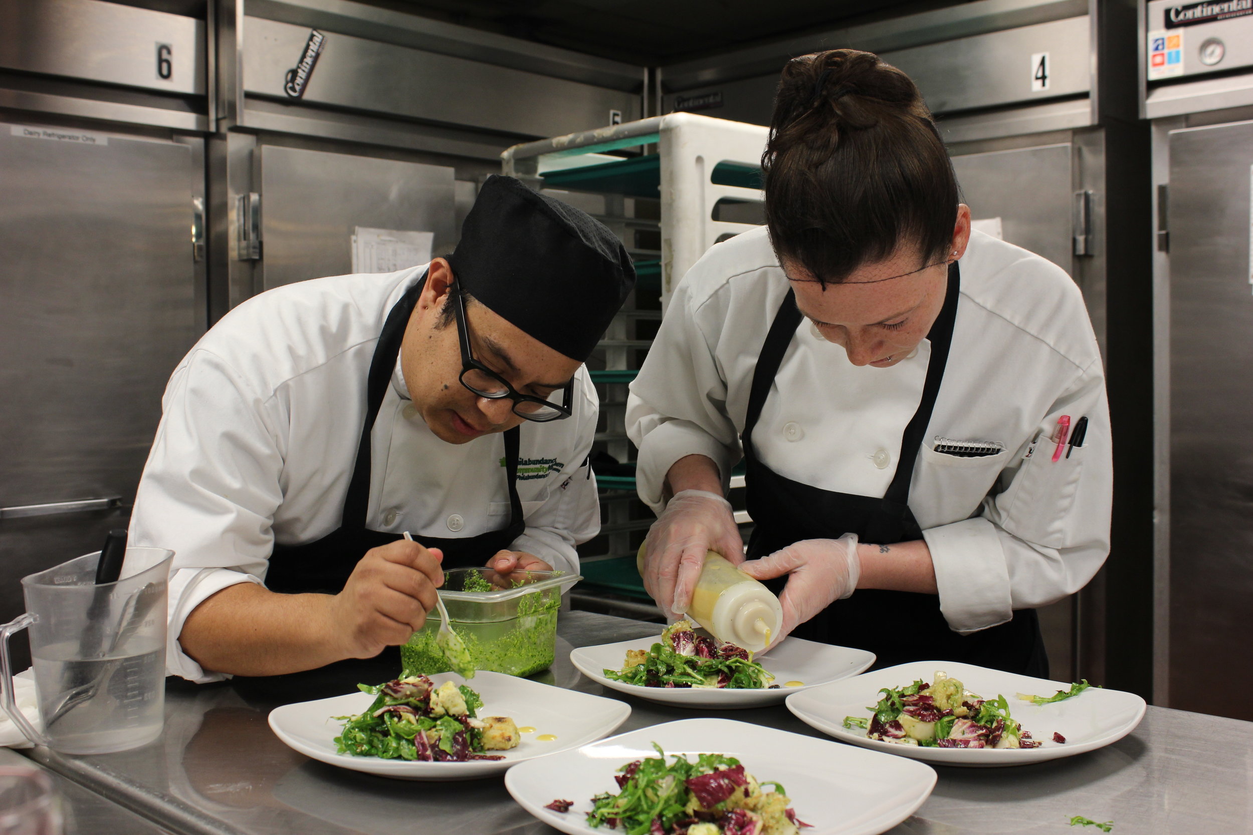 Alt text: two chefs in white jackets and black aprons lean over plates of salad while placing green dressing and vinegar onto them.