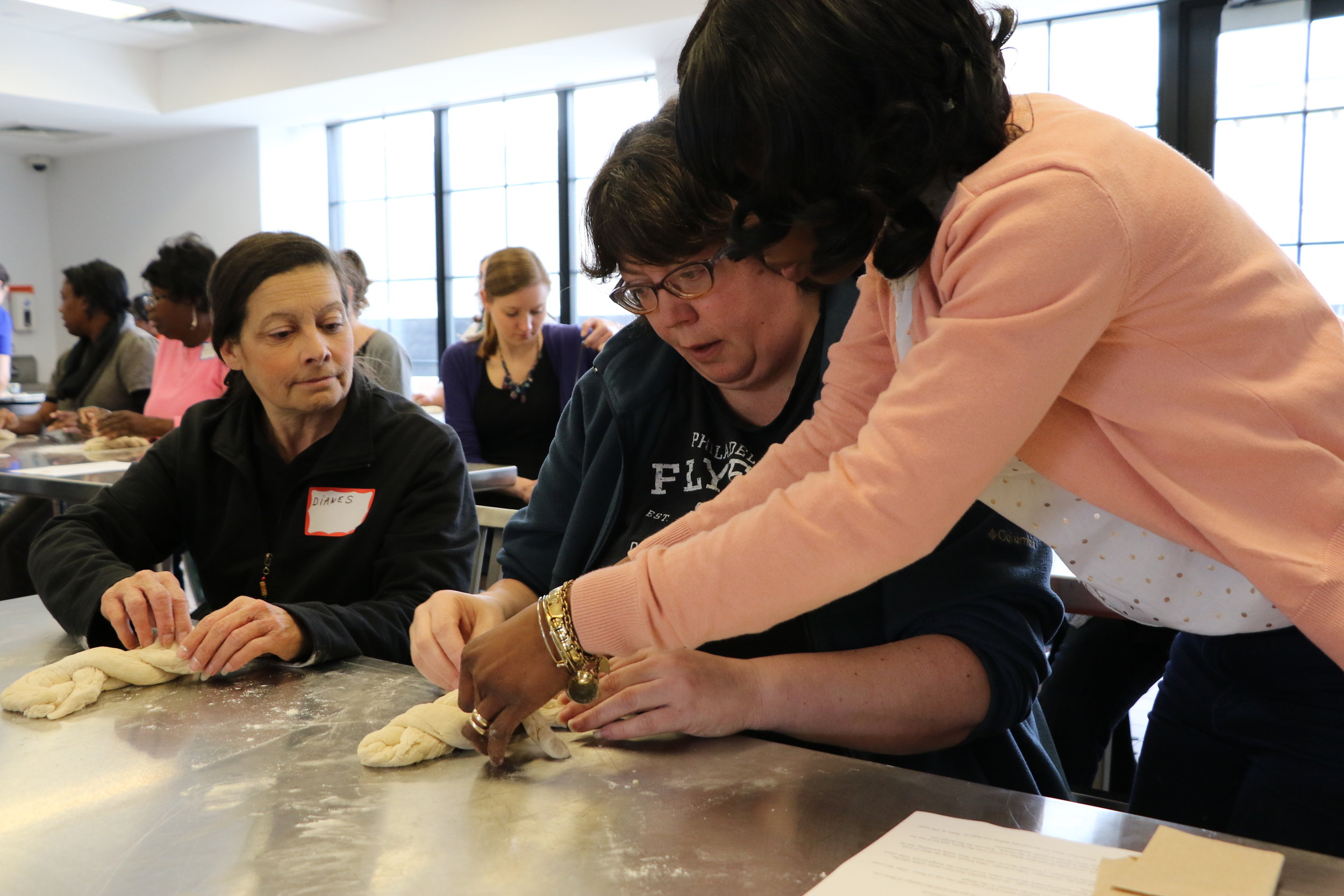 Alt text: three women work together at a stainless steel table to knead dough into a braid.