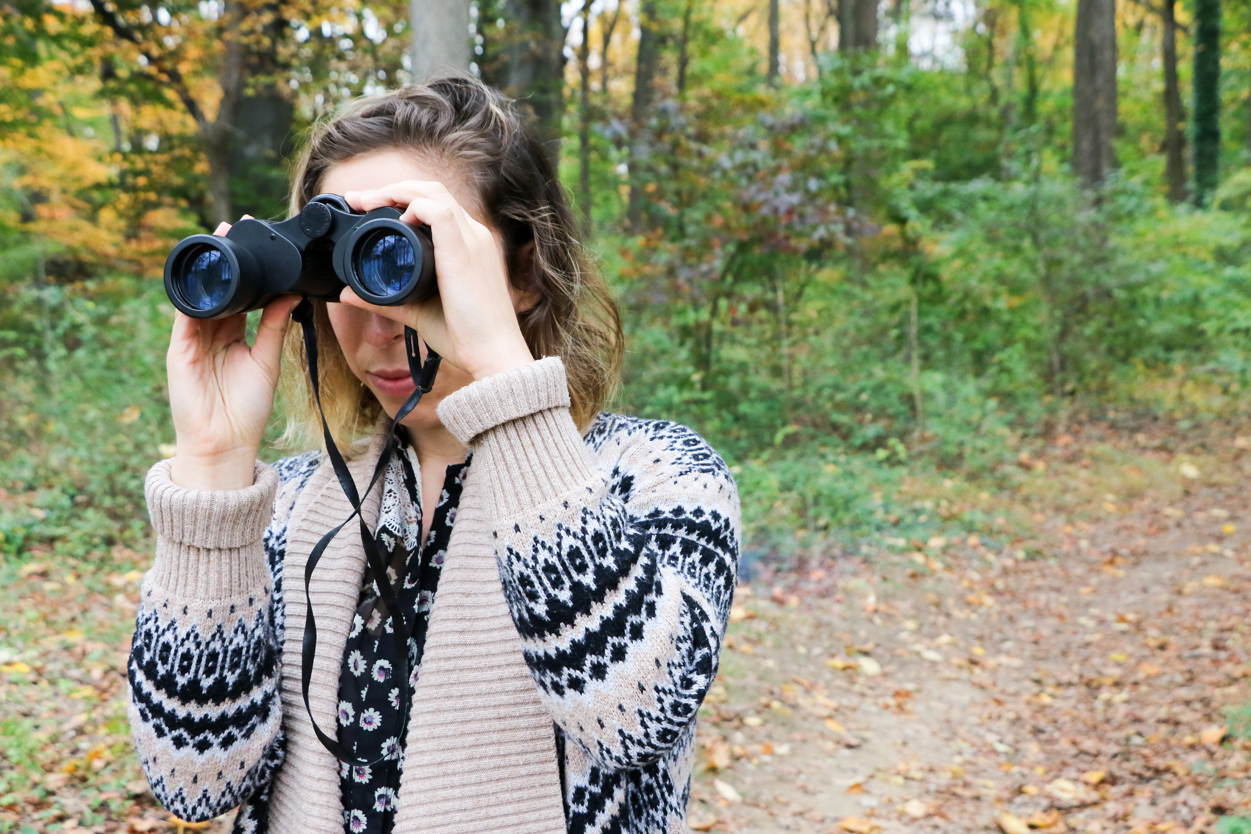 Alt text:  a young white woman with streaky brown and blonde hair looks through big black binoculars in the middle of a nature path. She’s wearing a thick blue and tan cardigan over a floral shirt.