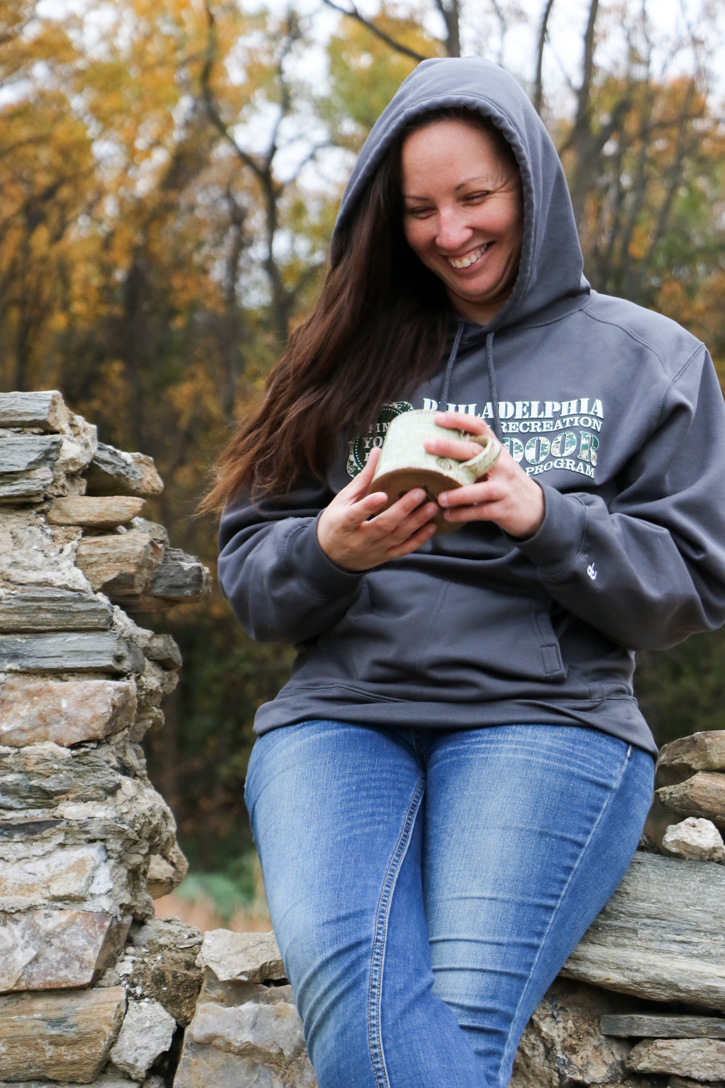 Alt text: a young white woman with long brown hair leans against a gray rock formation while holding a cup of hot tea. She’s smiling while looking down at the tea and wearing a gray and green Philadel