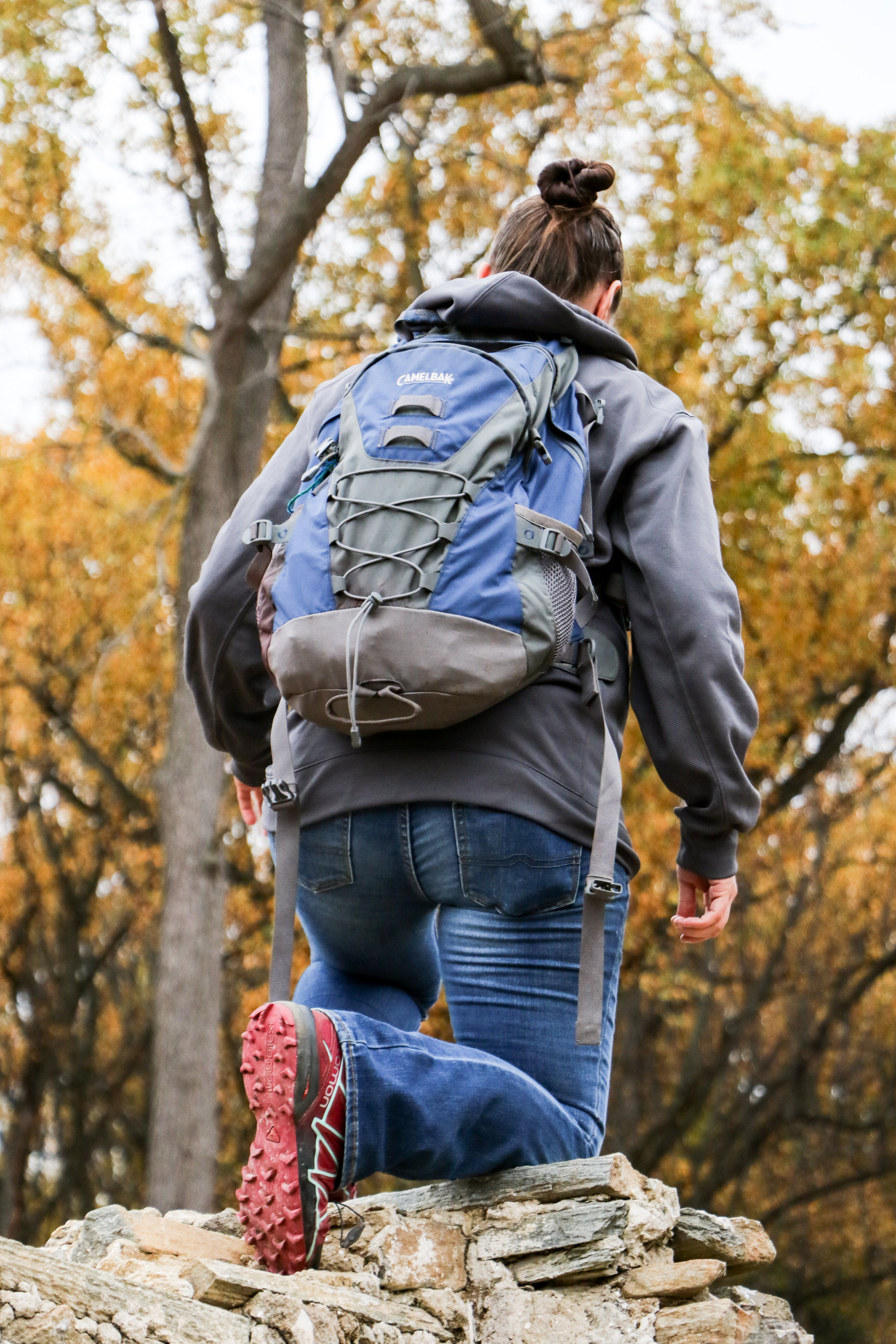 Alt text: a person with their back to the camera steps up onto a gray rock formation. They’re wearing a strappy blue backpack over a gray sweatshirt and blue jeans with hiking boots. The bottom of the