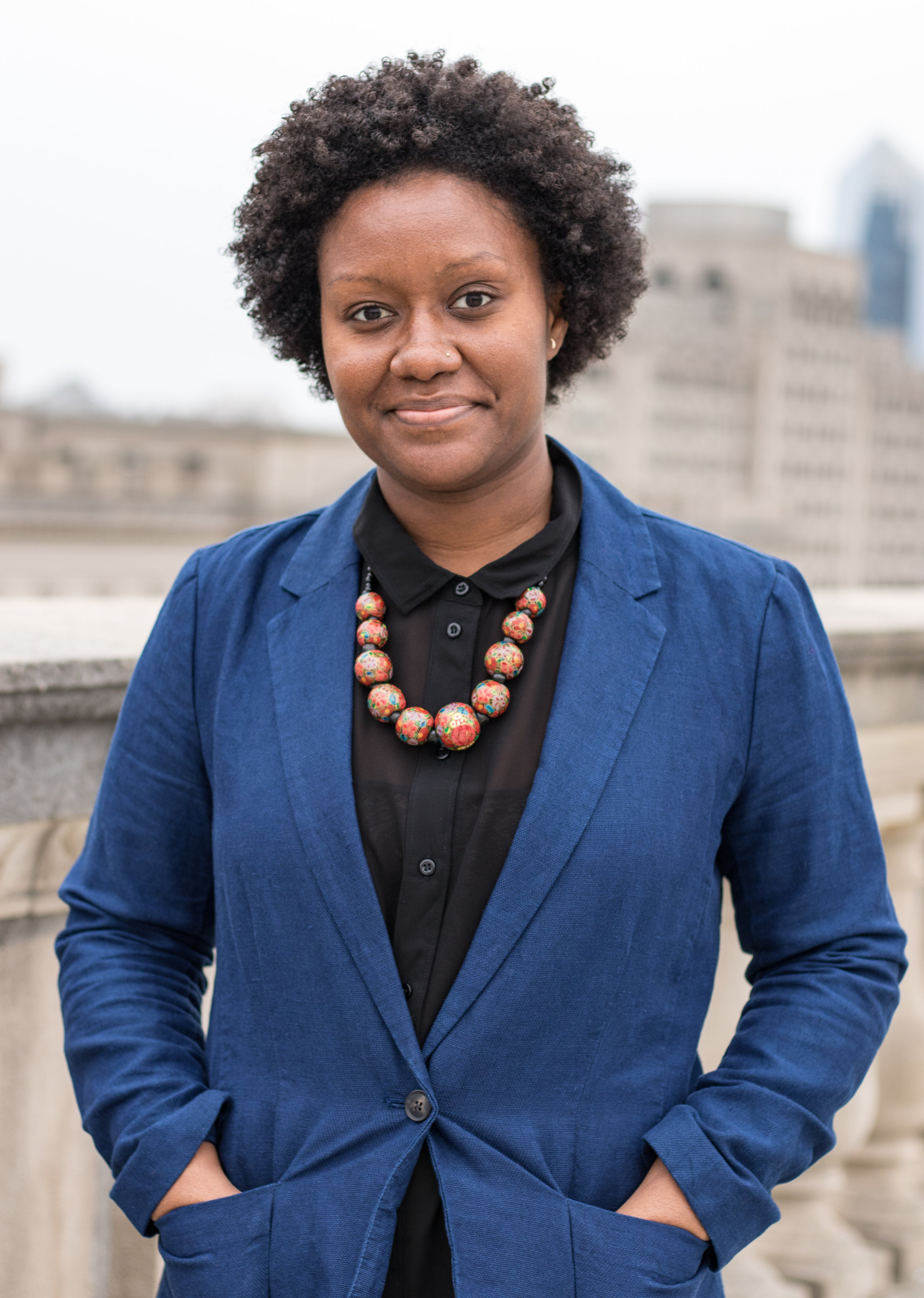 Alt text: a young black woman smiles at camera while wearing a blue blazer and black blouse along with a chunky, rounded wooden bead necklace. Her hands are in the blazer pockets. The background is bl