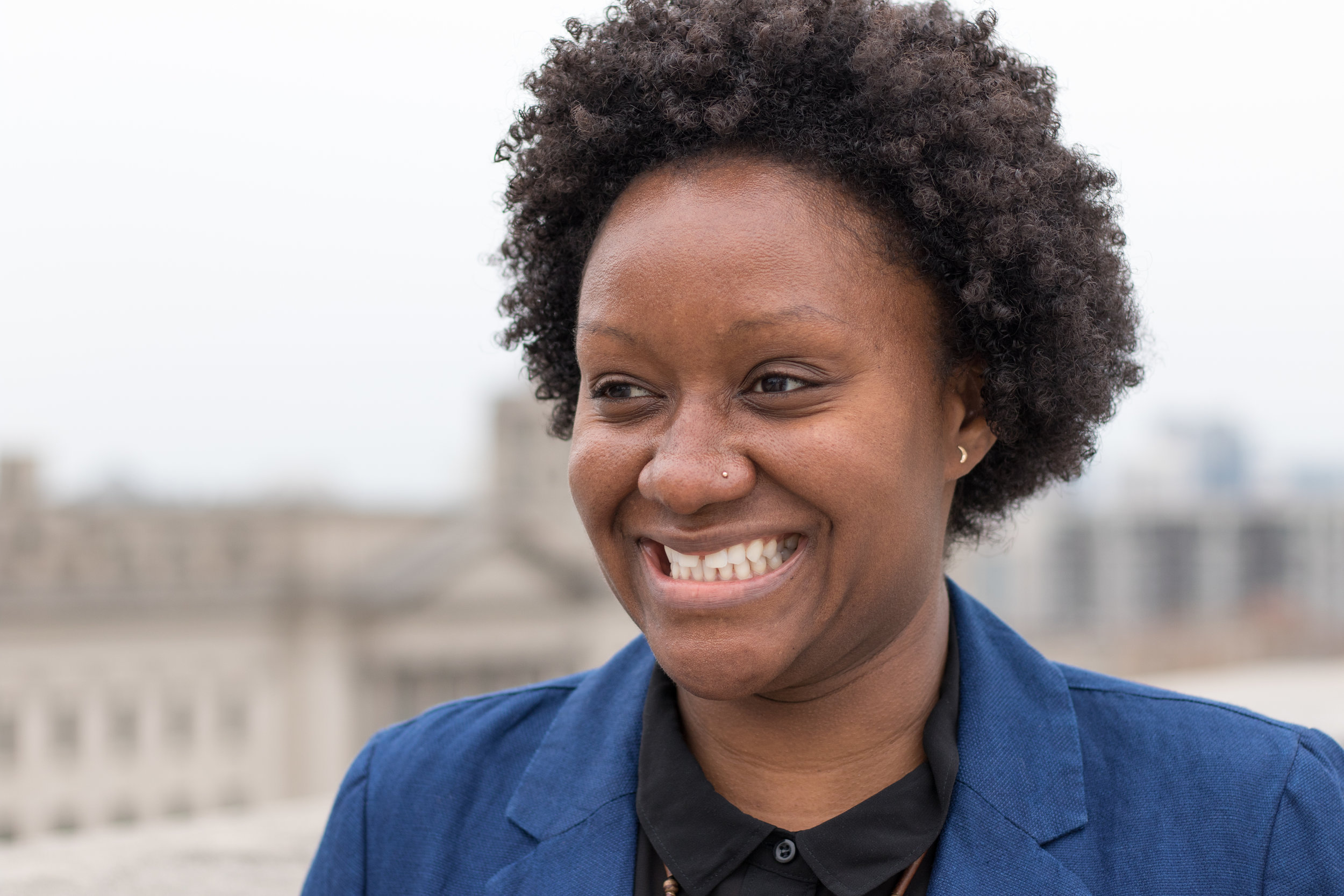 Alt text: a young black woman smiles a gap-toothed grin while looking off camera and wearing a blue blazer and black blouse. The background is blurry rooftops of buildings.