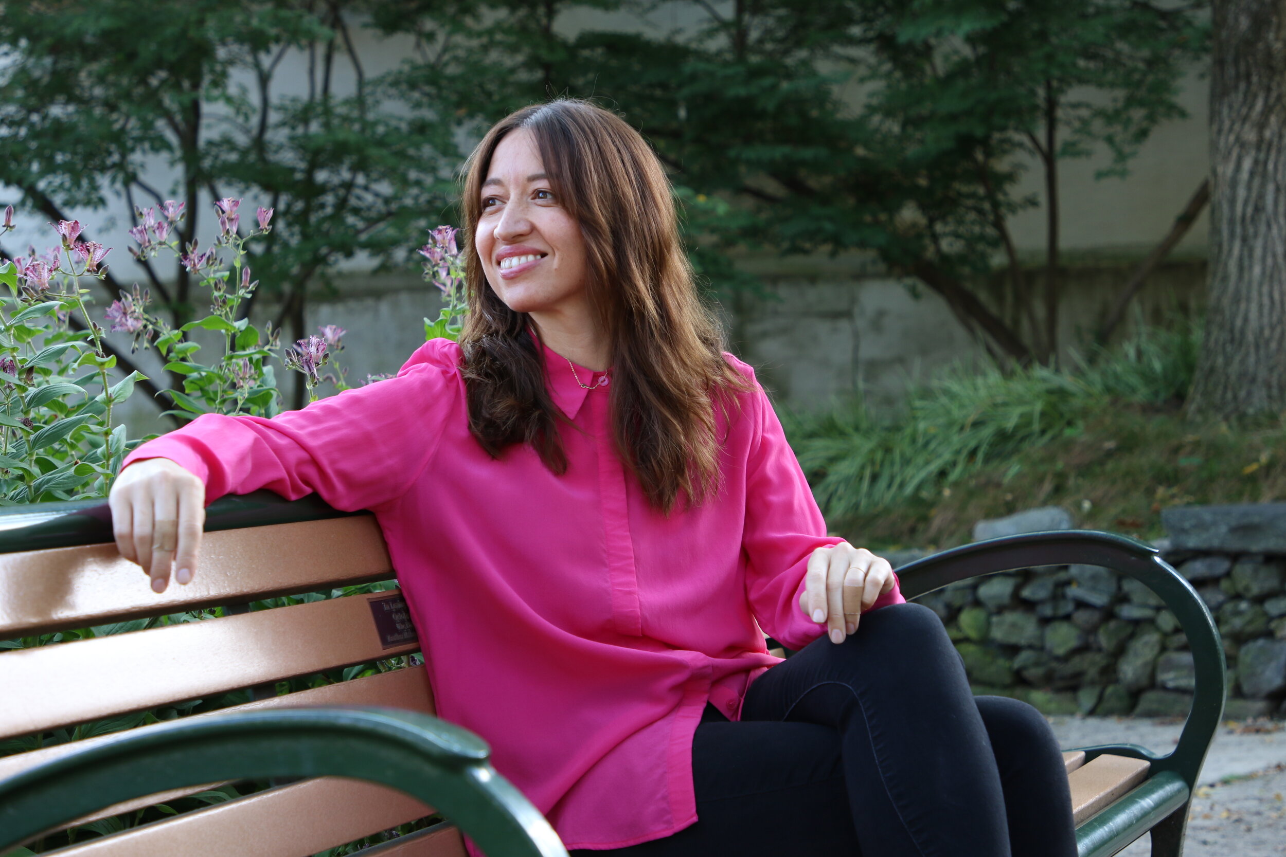 Alt text: a young asian american woman sits on a park bench and smiles while looking off camera. She has medium-length brown hair over her shoulders and is wearing a fuschia button down blouse with bl