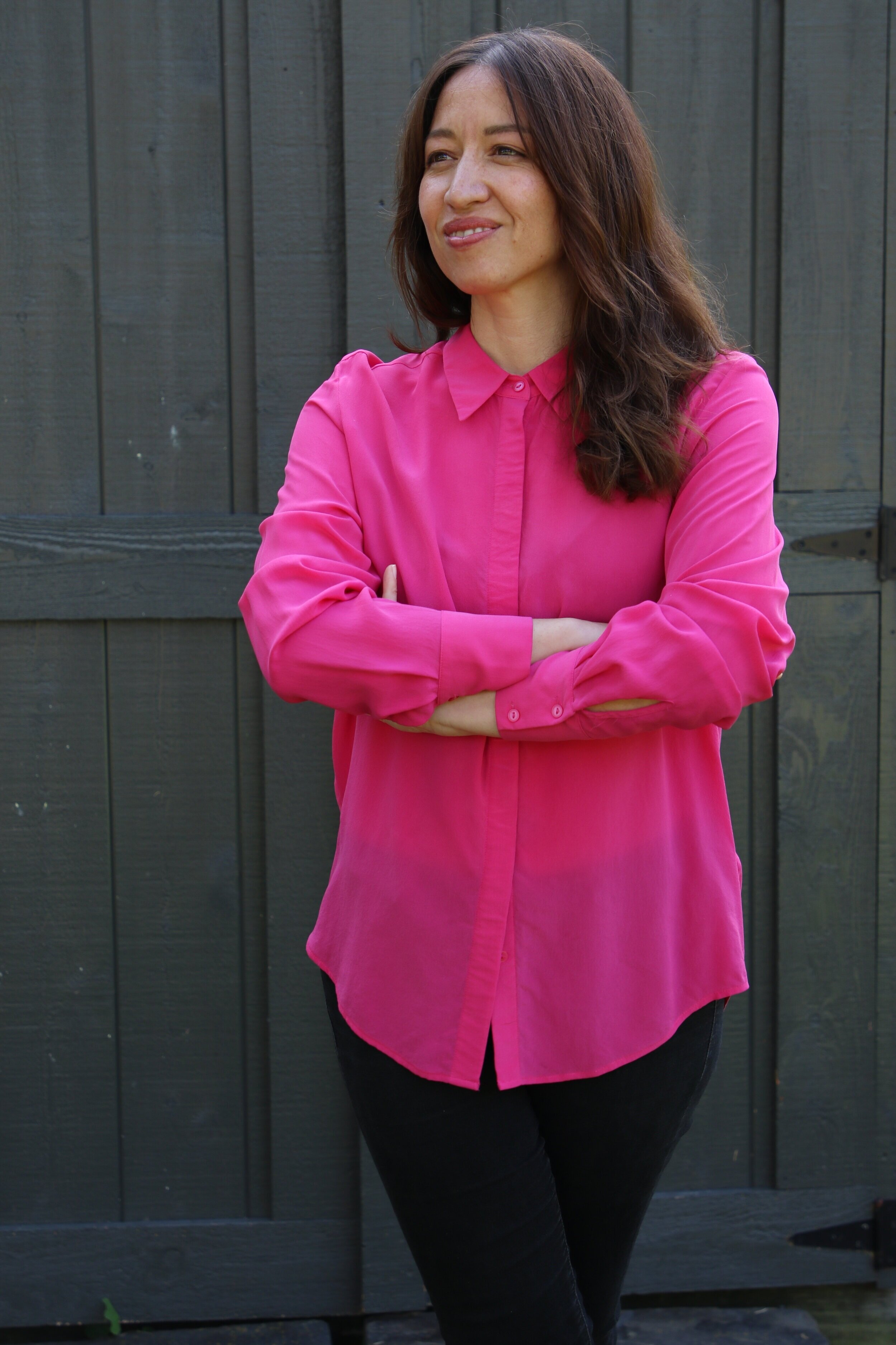 Alt text: a young asian american woman smiles confidently while looking off camera. She has medium-length brown hair over her shoulders and is wearing a fuschia button down blouse with black pants. He