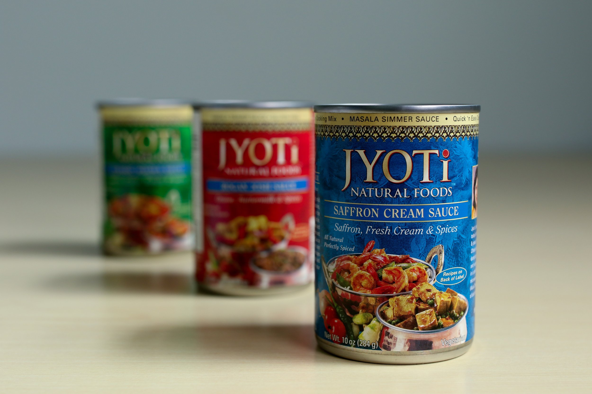 Alt text: a blue, red, and green can of indian sauce sit in a diagonal row. The blue can at the front reads “jyoti natural foods saffron cream sauce”