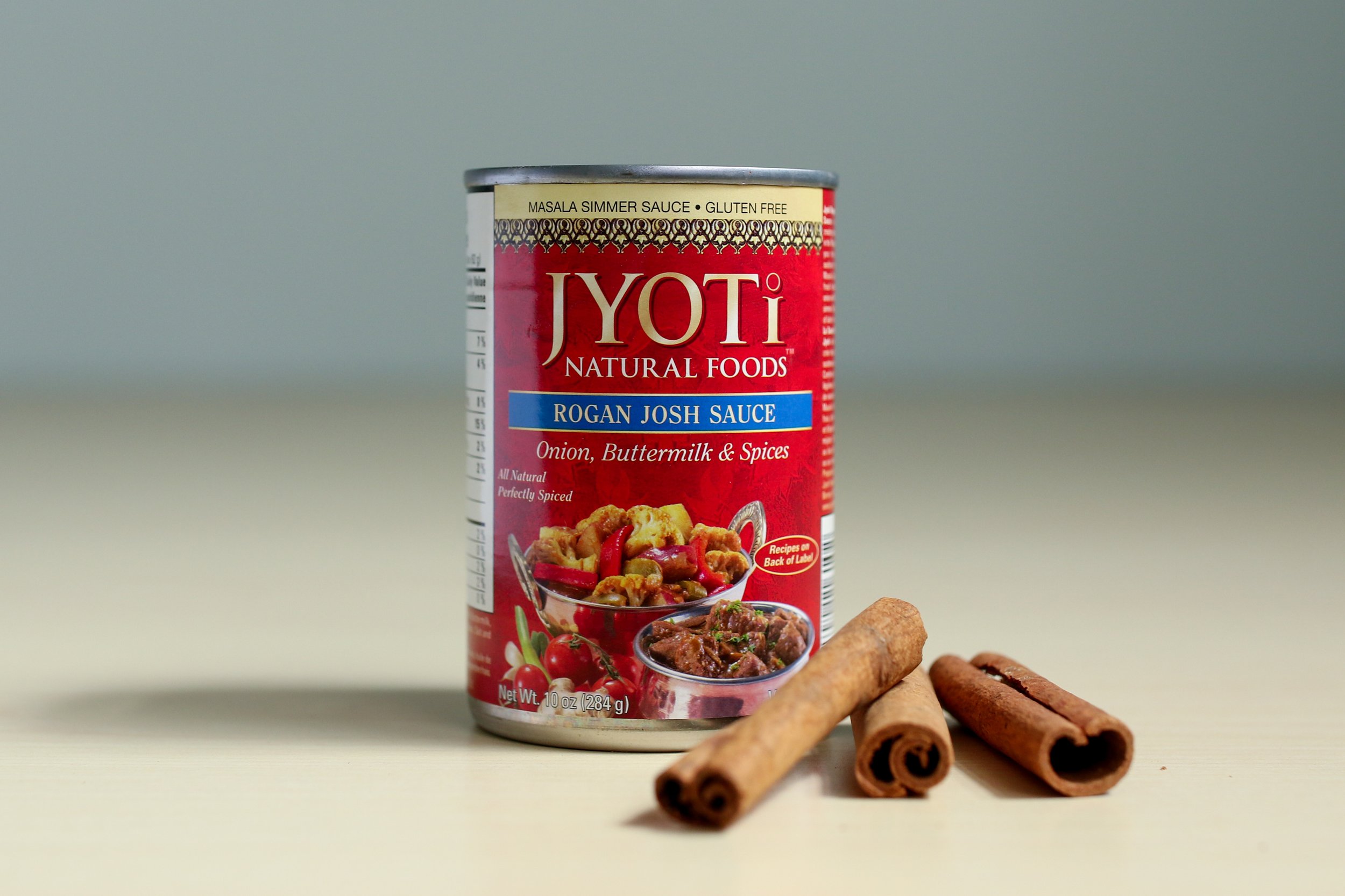 Alt text: a bright red can of indian sauce reads “jyoti natural foods rogan josh sauce” with three sticks of cinnamon lean against its side.
