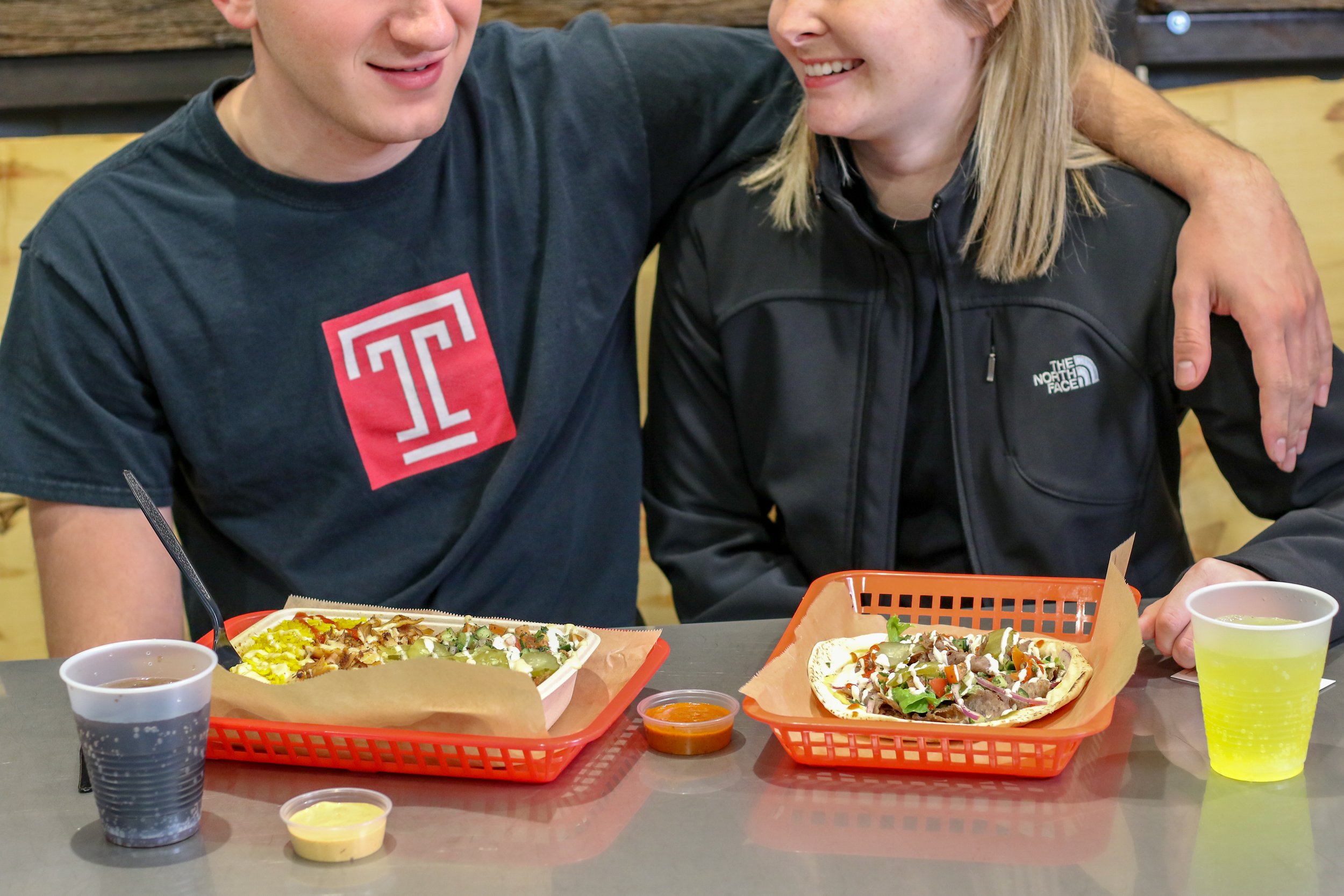 Alt text: two young white students sit together at lunch. One person wears a black and red temple university tshirt and drapes his arm over the other person who is wearing a black fleece jacket. In fr