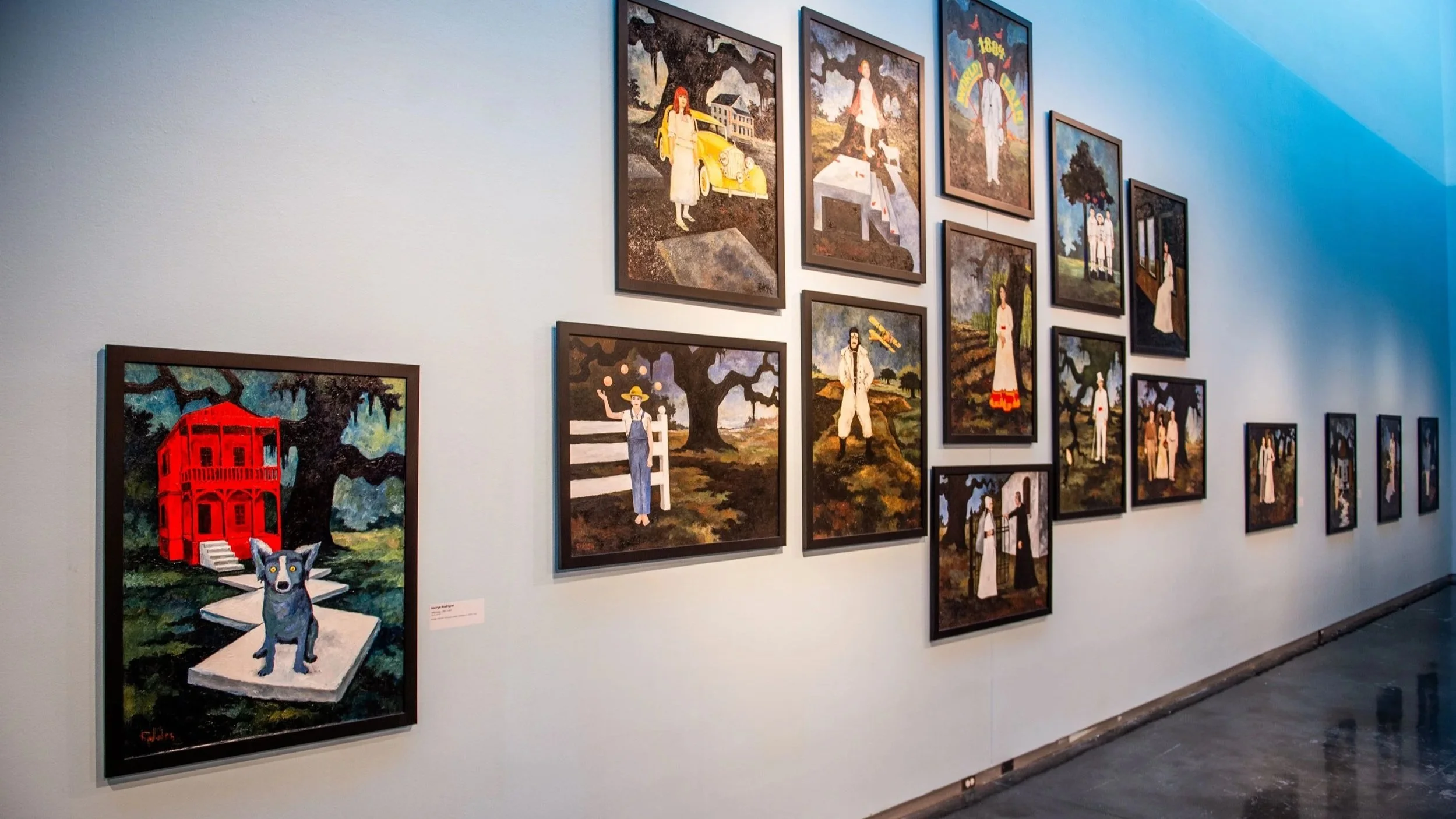 Gallery wall with multiple framed paintings arranged salon-style, depicting figures in outdoor scenes; at left, a painting shows a red house with a blue dog in the foreground.