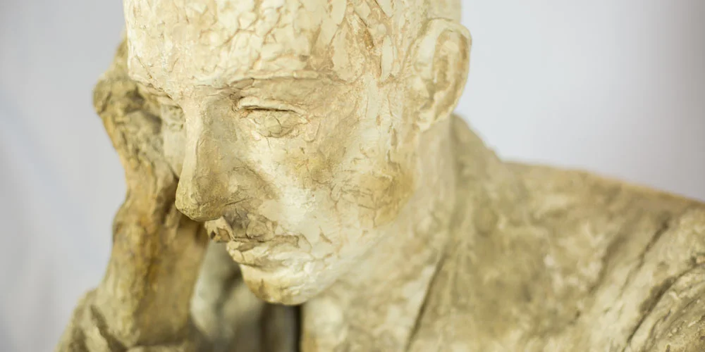 Close-up of a gold-toned sculpture by Angela Gregory depicting a contemplative figure with head resting on one hand, showing detailed facial features and textured surface.