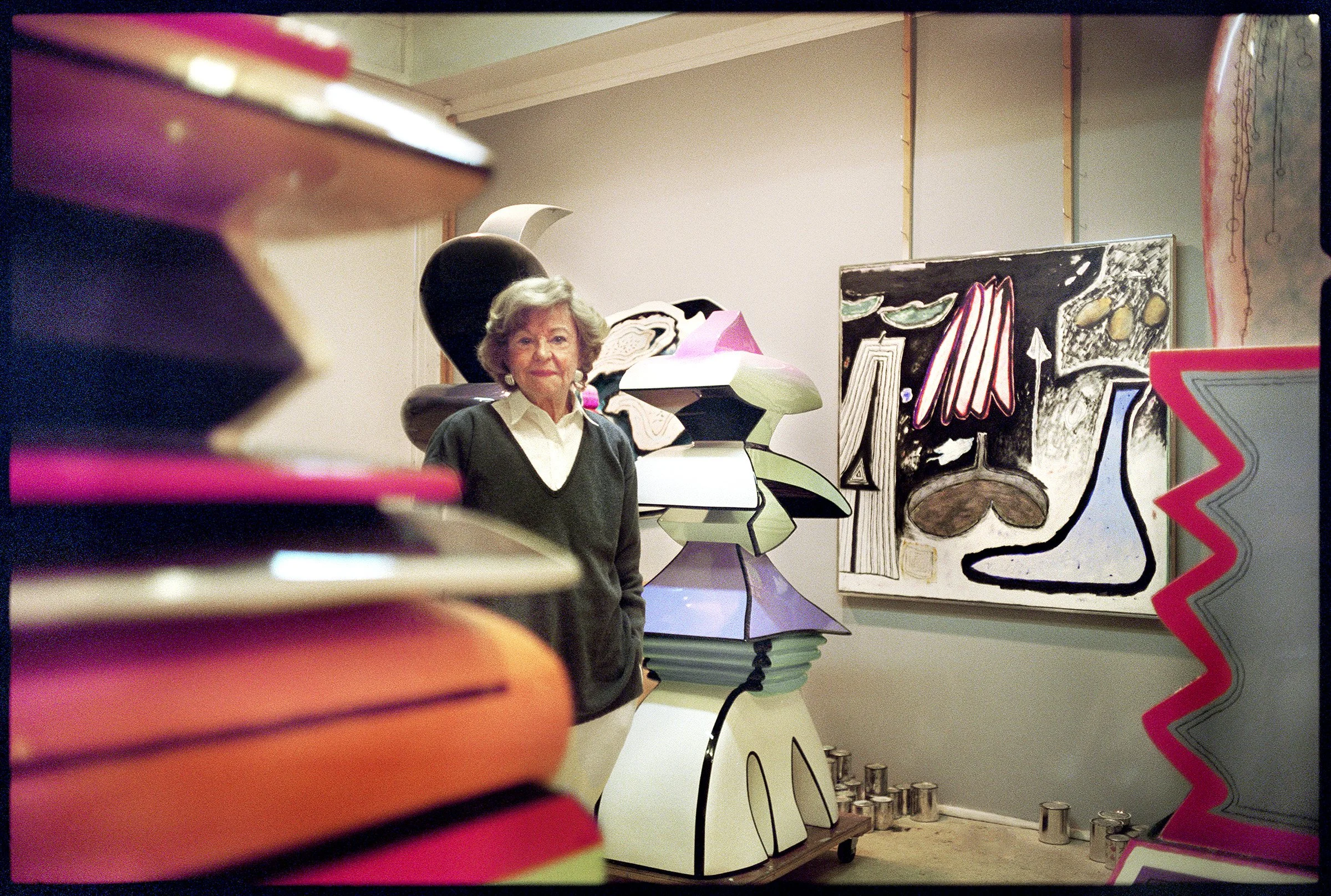 artist Ida Kohlmeyer stands in a studio among colorful sculptural forms and a large painting.