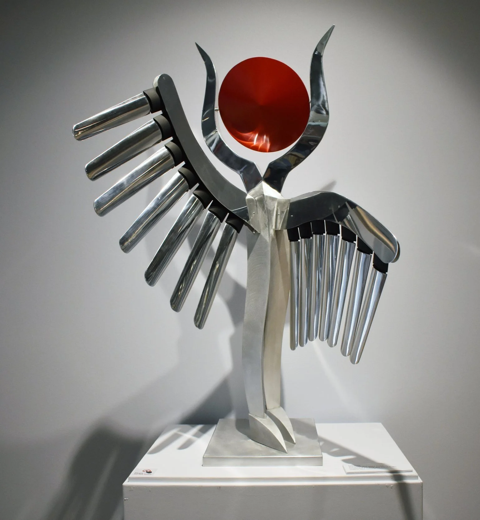 a silver winged sculpture with tubular elements and a red circular disk by artist Lin Emery.