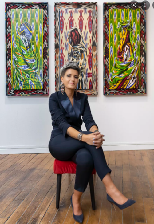 Portrait of artist Alia Ali seated on a red chair in a gallery, wearing a dark suit and heels, with colorful patterned artworks displayed on the wall behind her.