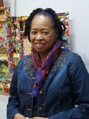 Portrait of artist Malaika Favorite standing in front of a colorful painting, wearing a denim jacket and a patterned scarf, and smiling toward the camera.