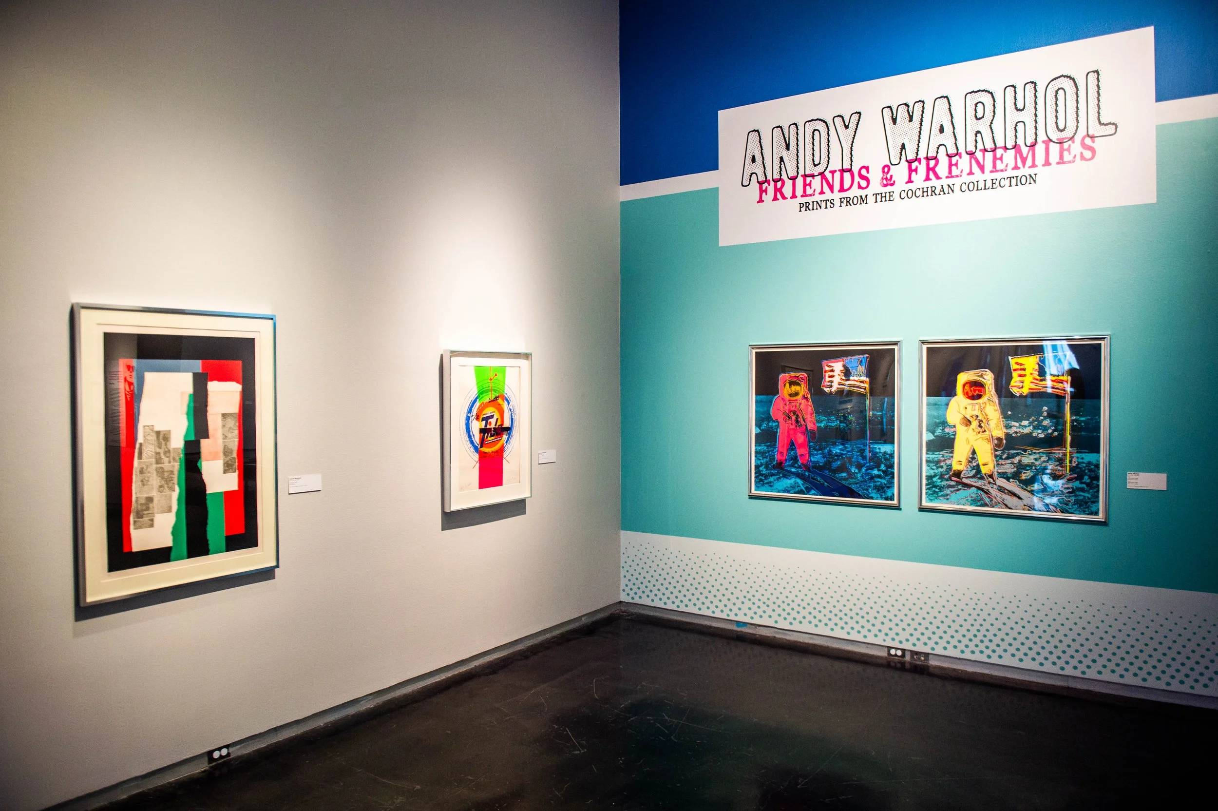 Gallery corner with wall text reading “Andy Warhol: Friends & Frenemies,” featuring framed artworks on white and blue walls, including colorful prints of figures in space suits.