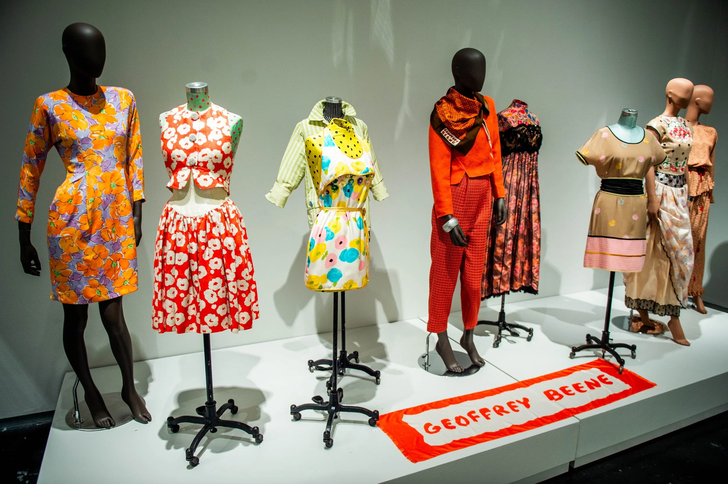 Display of mannequins wearing colorful patterned garments, including dresses and coordinated outfits, arranged on a platform with a textile reading “Geoffrey Beene” in front.