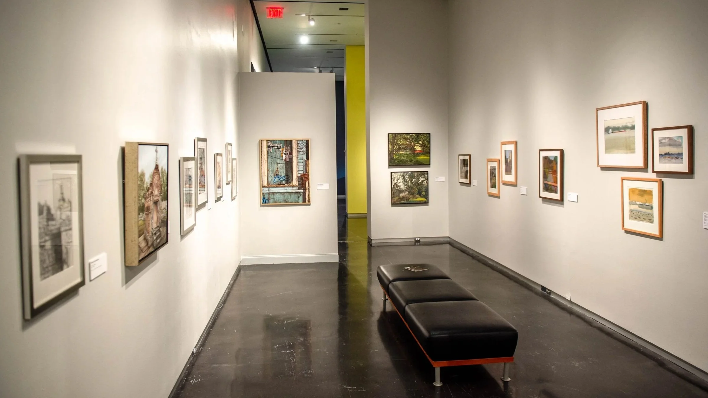 Narrow gallery corridor with framed artworks lining both walls and a black bench centered on the floor, leading toward additional gallery space in the distance.