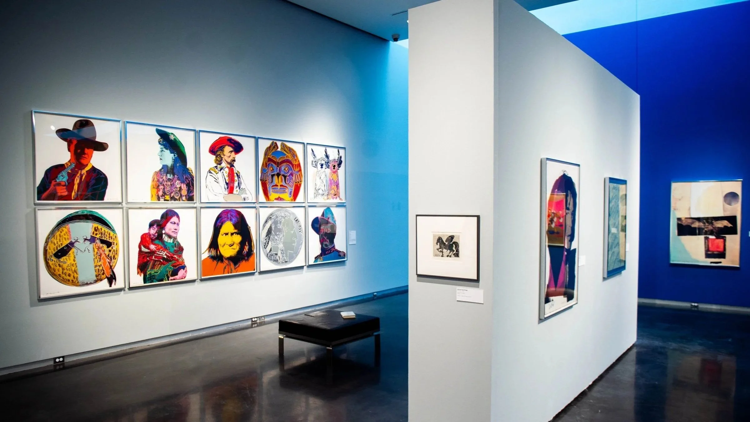 Gallery view with a grid of colorful portrait prints on one wall, additional framed works by artist Andy Warhol on adjacent walls, and a bench centered in the space.