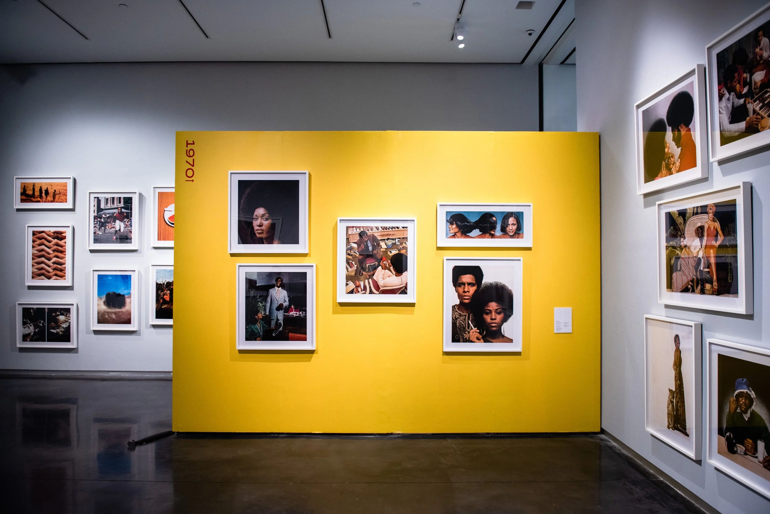 Gallery view with framed portrait photographs displayed on a large yellow wall labeled “1970,” with additional photographs visible on surrounding walls.