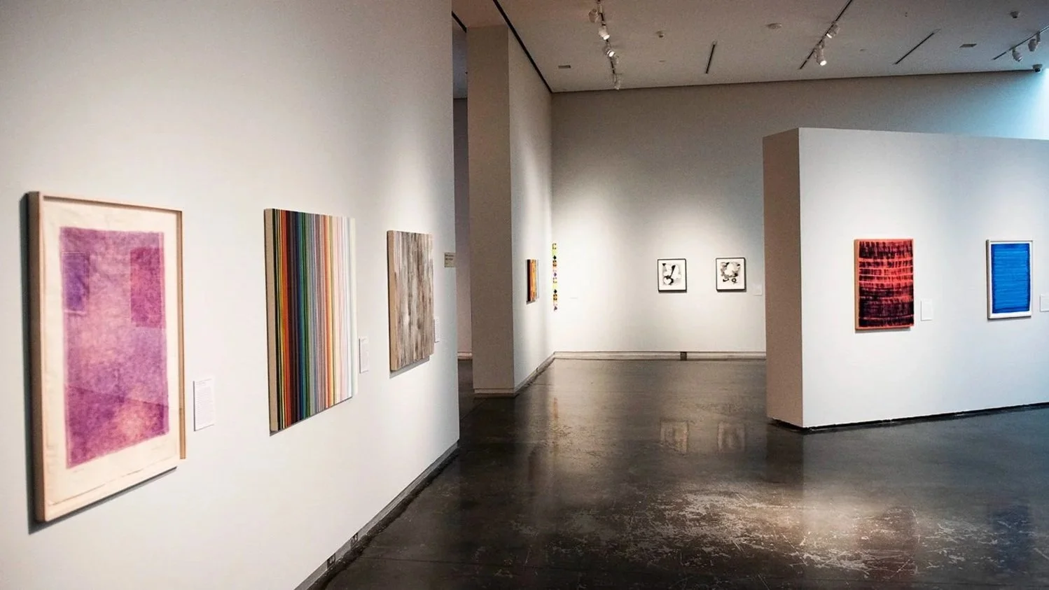 Wide gallery view with contemporary artworks on white walls, including abstract paintings and prints, arranged around a central partition with open floor space and overhead track lighting.