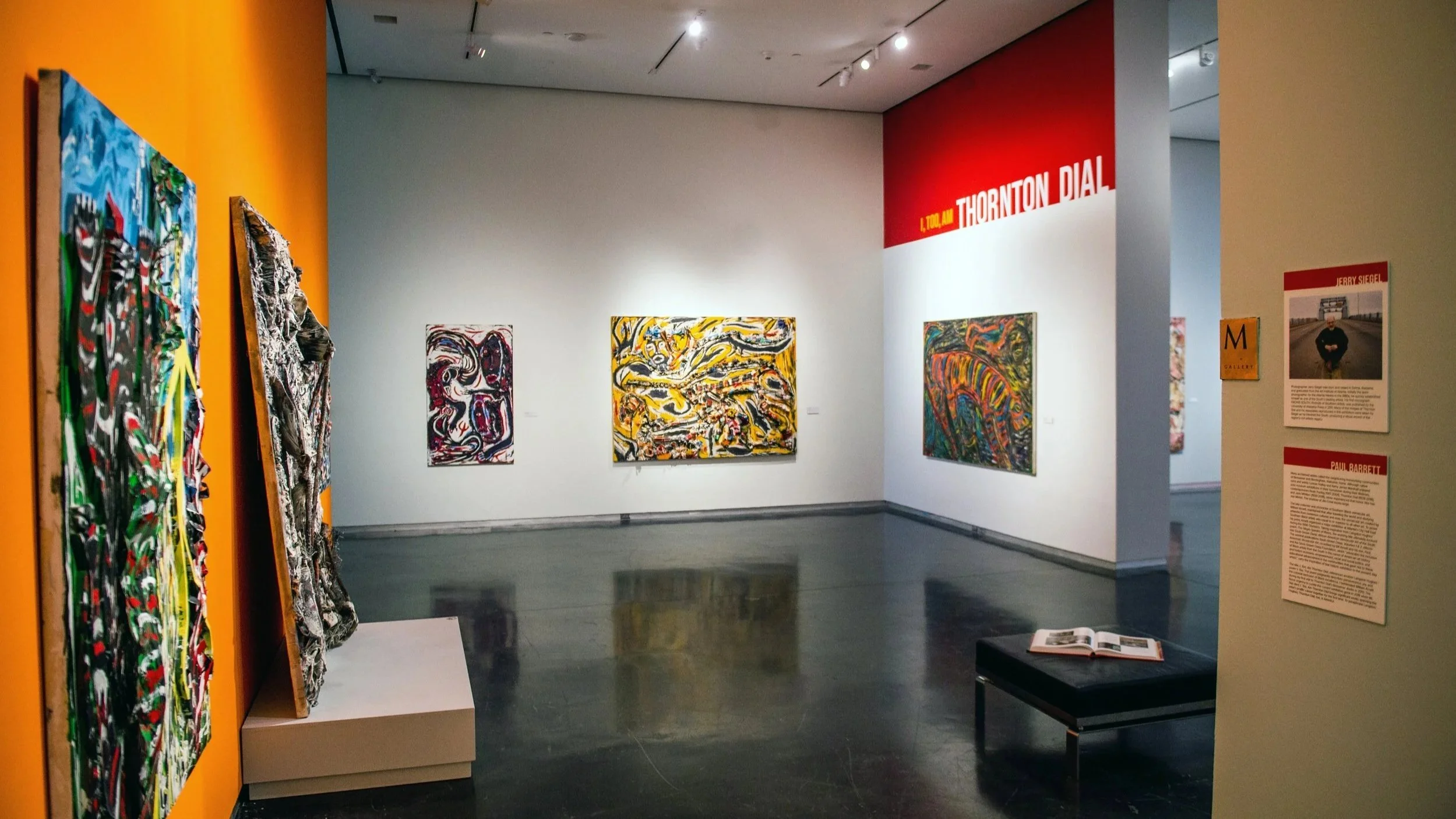 Gallery view featuring paintings and sculptural works, with wall text reading “Thornton Dial,” and a bench with an open book in the foreground.
