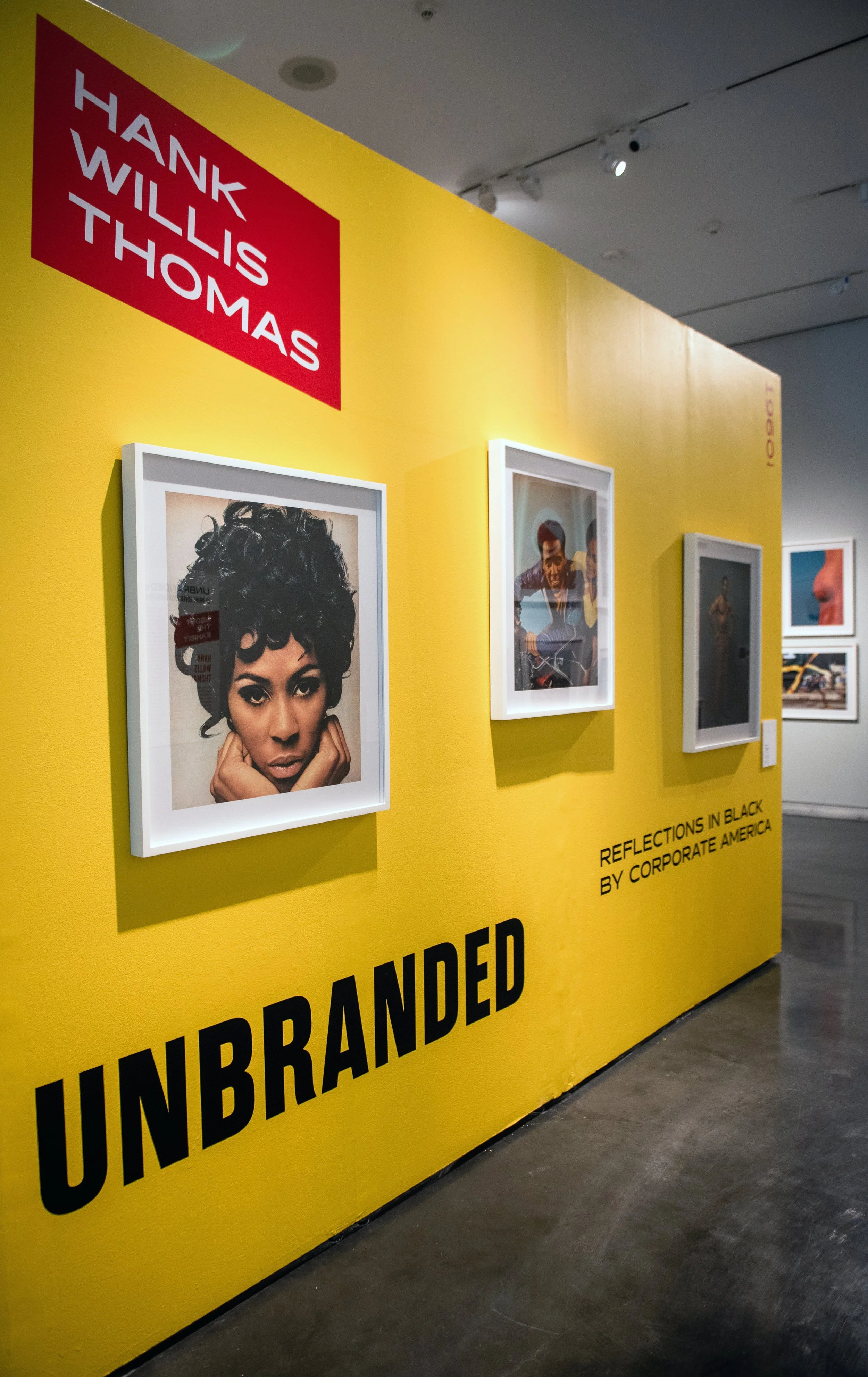 Gallery wall with yellow backdrop reading “Hank Willis Thomas: Unbranded,” featuring framed photographic portraits and additional works extending along the wall.
