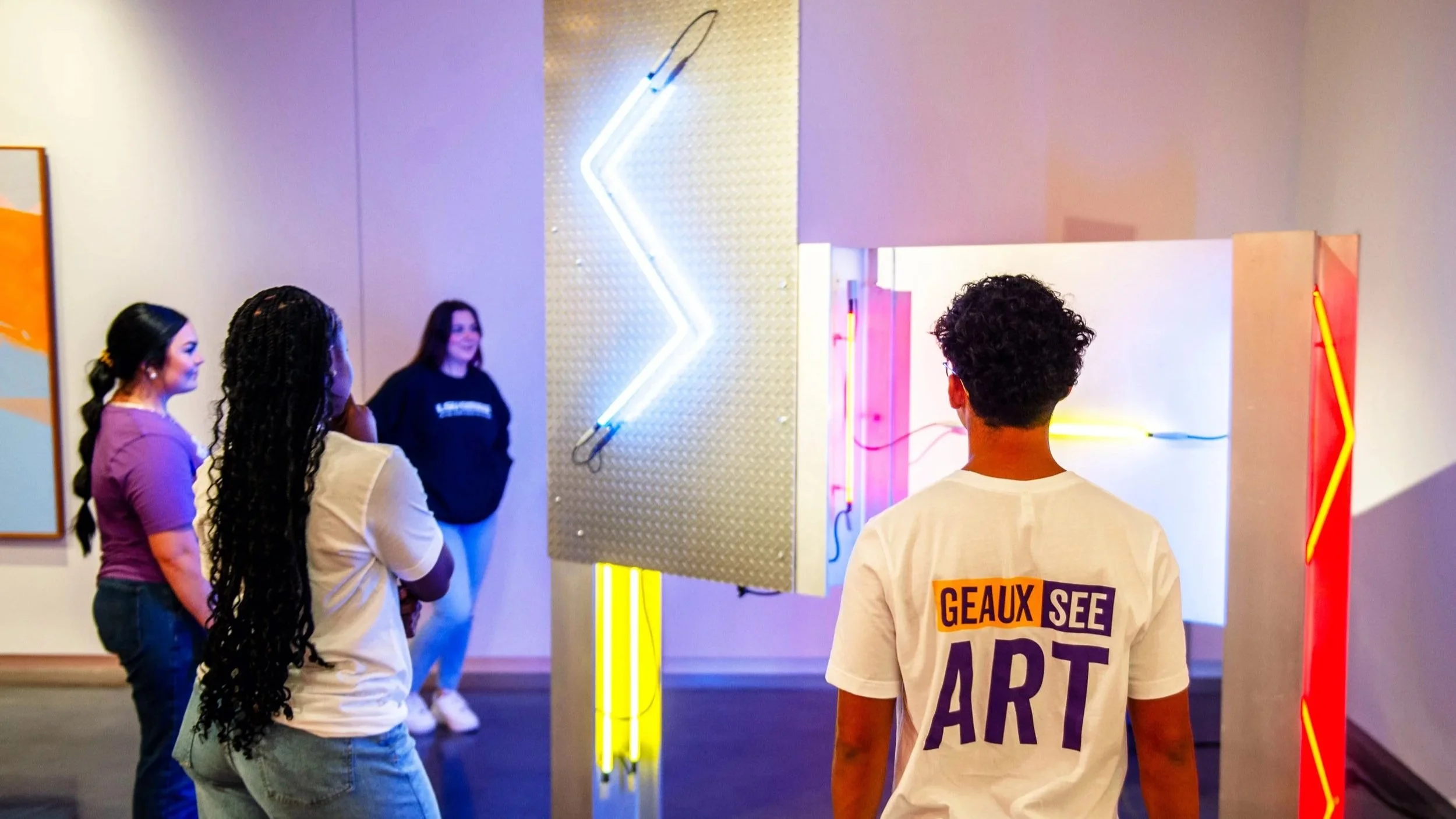 Visitors stand in a gallery viewing a neon light installation; a person in the foreground wears a shirt reading “Geaux See Art” while others observe the artwork.