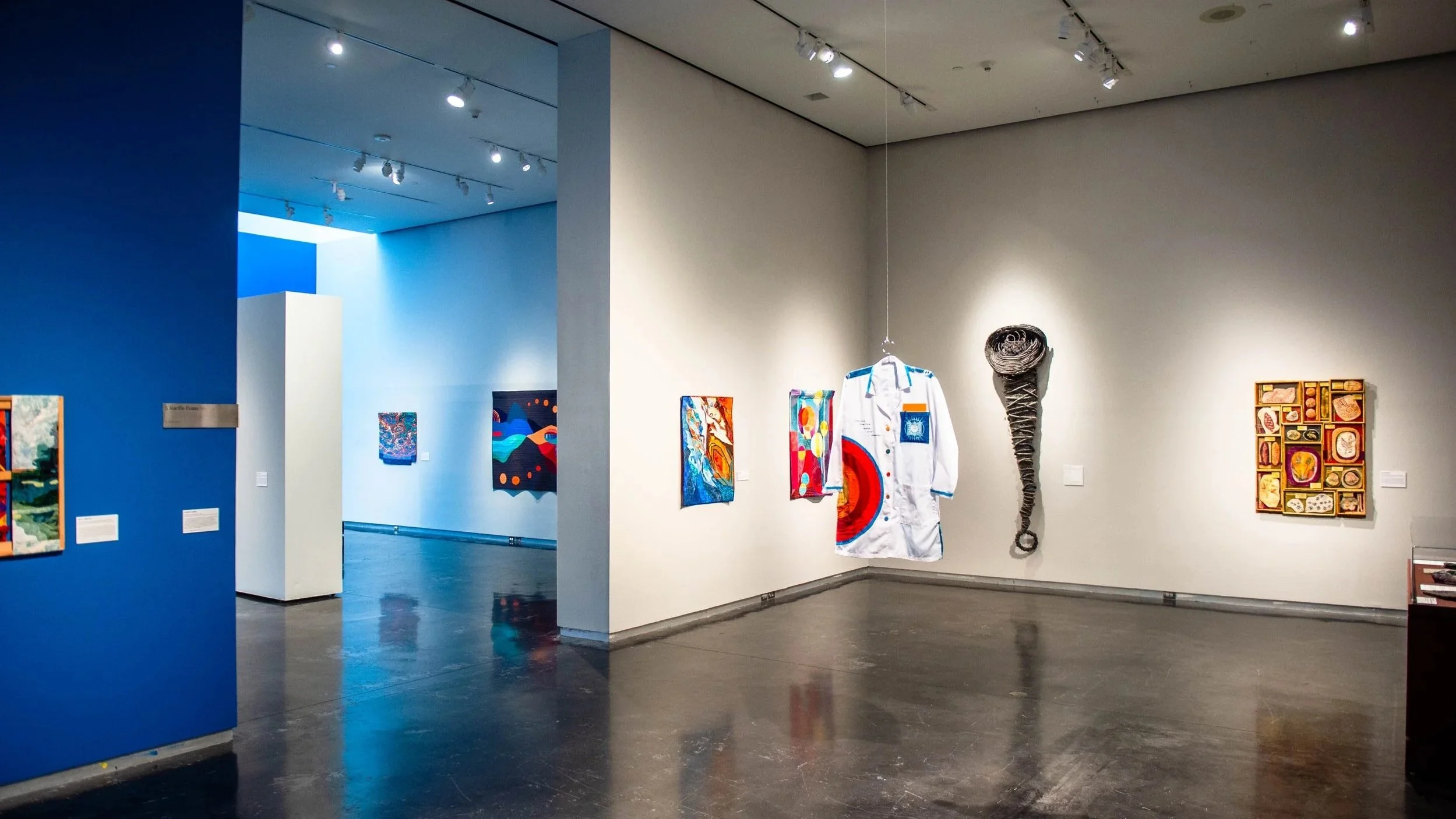 Gallery view with contemporary artworks on white walls, including a hanging shirt artwork, a coiled sculptural piece, and colorful paintings, with additional works visible in an adjacent blue-walled gallery.