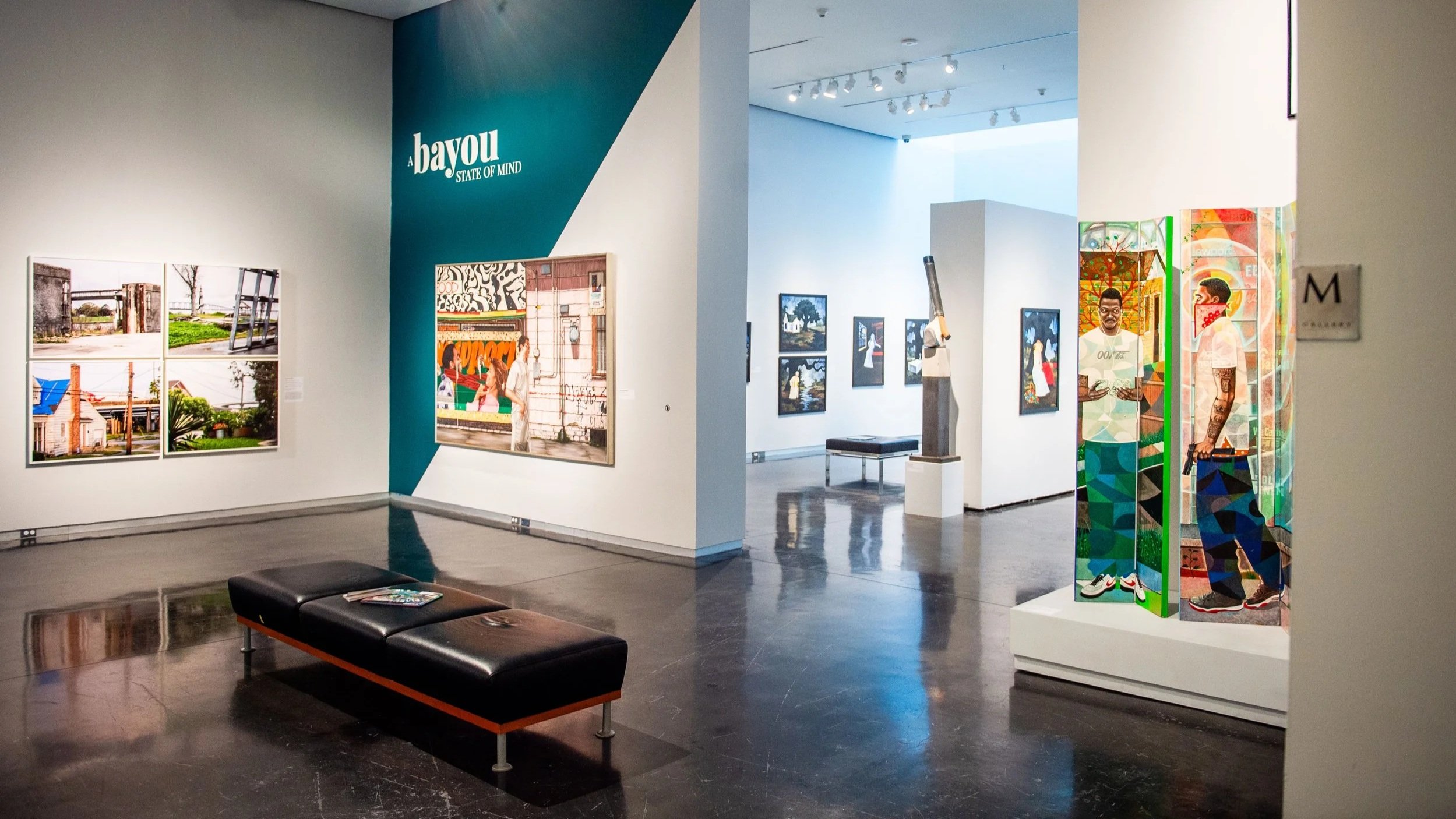 Gallery view featuring the exhibition “A Bayou State of Mind,” with paintings on the walls, a bench in the center, and a colorful multi-panel artwork on a pedestal in the foreground.