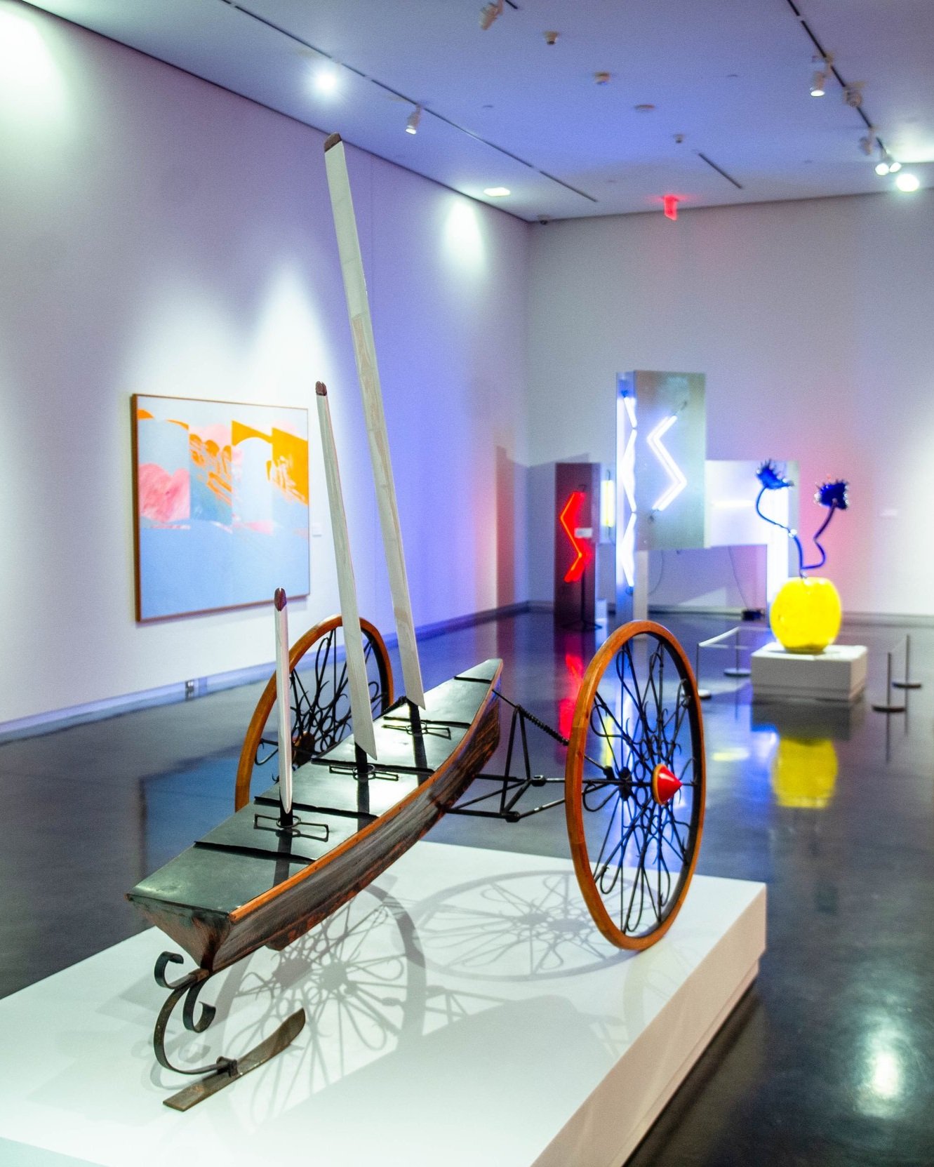 This is a gallery view of the LSU Museum of Art's modern contemporary gallery featuring a large sailboat on wheels, paintings, and multicolor neon sculpture, and a yellow vase glass sculpture in the room.