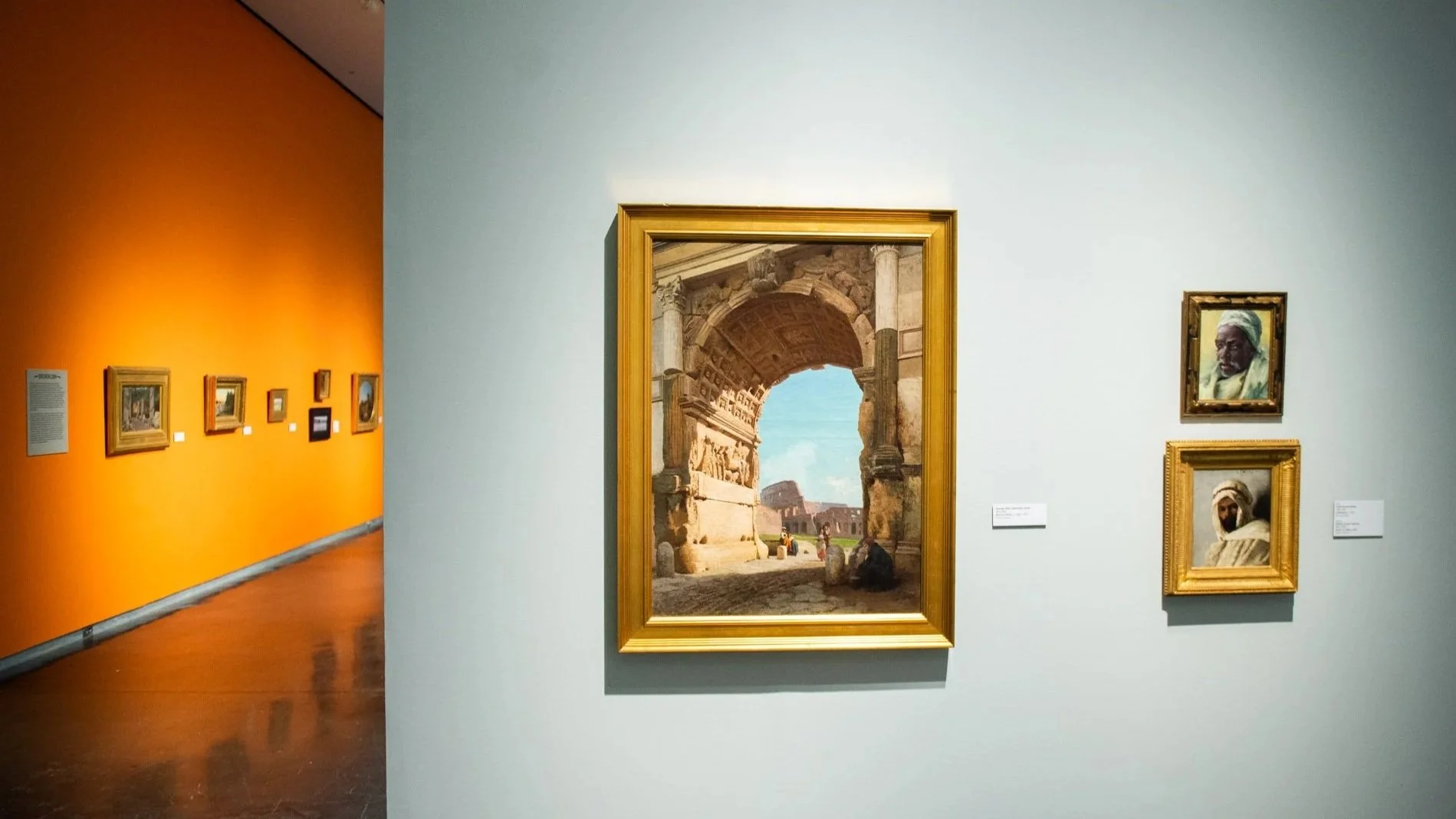 Gallery view featuring a large framed painting of a classical stone archway opening onto a sunlit scene, with two smaller portrait paintings to the right and additional works visible along an adjacent orange wall.
