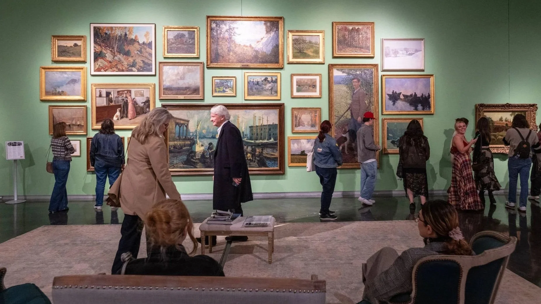 Visitors explore a gallery with a green wall densely hung with framed paintings, while others stand, walk, or sit on nearby seating viewing the artworks.